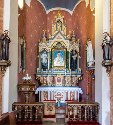 Zdjęcie nr 1: Ołtarz przyścienny, architektoniczny, jednokondygnacyjny, trójosiowy, ze zwieńczeniem. Ołtarz prostopadłościenny z antepedium artykułowanym trzema płycinami. Dwie skrajne, węższe zawierają dekorację złożoną z korony i nasadzonego na nią krzyża pomiędzy liśćmi wawrzynu ustawionymi w słup, środkowa, szersza z motywem krzyża równoramiennego o trójlistnych zakończeniach ramion, ujętego maswerkiem. Predella wklęśle zwężająca się, po bokach artykułowana pięcioma płycinami ze stylizowanymi wiciami roślinnymi. Tabernakulum w formie prostopadłościennej szafki, flankowane od frontu parą kolumienek wtopionych w naroża, zamknięte nadwieszonym łukiem półkolistym. Prostokątne, półkoliście zamknięte drzwiczki zdobione płaskorzeźbionym motywem kielicha eucharystycznego.  Część środkowa nastawy w formie trójprzelotowego łuku triumfalnego. Każda z ujmujących płytkie wnęki półkolistych arkad wsparta na parze kolumn. Podłucza ujęte dekoracyjnym ornamentem arkadkowym, który w części środkowej przybiera kształt trójliścia. Kolumny wspierają zamykające osie wimpergi. Zwieńczenie części środkowej retabulum osadzone na zdwojonym cokole, ma formę wspartej na parze kolumienek półkolistej arkady, której podłucze wypełnia motyw trójliścia, zamkniętej wimpergą. Całość ujmuje para przypór o zakończeniach ujętych kolumienkami wspierającymi sterczyny. Boczne osie zwieńczone motywem oktogonalnych latarni przekrytych kopułkami. Pola predelli zdobią motywy wici akantowych, boki ujmuje para ażurowych uszaków. W polach wimperg oraz w dolnej partii wnęki części środkowej stylizowana ornamentyka roślinna, w górnej części monogram maryjny. Krawędzie zwieńczenia zdobi motyw wici roślinnej, czołganki oraz sterczyny zwieńczone kwiatonami. We wnęce części środkowej ujęty rzeźbioną, złoconą ramą obraz Matki Boskiej z Dzieciątkiem. We wnęce po prawej stronie retabulum figura św. Stanisława Kostki, po lewej św. Alojzego Gonzagi. W zwieńczeniu figura anioła. Polichromia ołtarza utrzymana w kolorach beżu i różu, detale i ornamenty złocone. Kolumny środkowej osi zielone, marmoryzowane, gzymsy w przyczółkach w kolorach niebieskim i czerwonym.