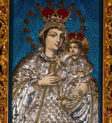 Zdjęcie nr 1: Obraz w kształcie stojącego prostokąta, ujęty rzeźbioną ramą z motywem wici akantowej, przedstawia Madonnę z Dzieciątkiem. Ukazana w półpostaci Maria zwrócona jest na wprost. W trzech czwartych skierowaną w lewo głowę charakteryzuje owalna twarz o wyrazistych rysach z ciemnymi łukami brwi, dużymi, migdałowatymi oczami o wyraźnych, dolnych powiekach, długim, prostym nosie, kształtnymi ustami w subtelnym uśmiechu i wyraźną linią podbródka. Partie szat pokryte są metalowymi osłonami. Maria ma na sobie srebrną, przewiązaną w pasie suknię zdobioną motywem tulipanów, pod szyją krótkie korale. Głowę, ramiona i ręce osłania srebrny, spięty pod szyją owalną broszą płaszcz obszyty złoconymi galonami, zdobiony motywem ukwieconej wici roślinnej. Matka Boska prawą, uniesioną na wysokości piersi ręką o wyprostowanej dłoni i złączonych trzech palcach wskazuje na trzymane na lewej ręce Dzieciątko. Zwrócony w trzech czwartych w prawo Jezus ukazany jest w pozycji siedzącej. Zaokrągloną twarz charakteryzują pełne, wyraziste rysy o dużych oczach z wydanie podkreślonymi liniami powiek, długim nosie, kształtnych ustach w delikatnym uśmiechu, zaróżowionych policzkach oraz wyraźniej linii podbródka. Jezus ma na sobie długą, srebrną, przewiązaną w pasie, zdobioną ukwieconą wicią roślinną suknię o złoconym obszyciu dekoltu i dekoracyjnych galonach przy mankietach dolnej krawędzi. Lewą ręką podtrzymuje małą, zamkniętą, złoconą księgę, prawą unosi w geście błogosławieństwa. Zwieńczone zamkniętymi, kameryzowanymi koronami głowy okalają – promienisty nimb Jezusa, wieniec z 12 gwiazd Marii. Tło niebieskie, z drobnym deseniem. 
