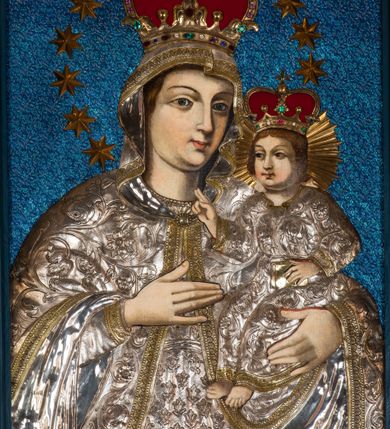 Zdjęcie nr 2: Obraz w kształcie stojącego prostokąta, ujęty rzeźbioną ramą z motywem wici akantowej, przedstawia Madonnę z Dzieciątkiem. Ukazana w półpostaci Maria zwrócona jest na wprost. W trzech czwartych skierowaną w lewo głowę charakteryzuje owalna twarz o wyrazistych rysach z ciemnymi łukami brwi, dużymi, migdałowatymi oczami o wyraźnych, dolnych powiekach, długim, prostym nosie, kształtnymi ustami w subtelnym uśmiechu i wyraźną linią podbródka. Partie szat pokryte są metalowymi osłonami. Maria ma na sobie srebrną, przewiązaną w pasie suknię zdobioną motywem tulipanów, pod szyją krótkie korale. Głowę, ramiona i ręce osłania srebrny, spięty pod szyją owalną broszą płaszcz obszyty złoconymi galonami, zdobiony motywem ukwieconej wici roślinnej. Matka Boska prawą, uniesioną na wysokości piersi ręką o wyprostowanej dłoni i złączonych trzech palcach wskazuje na trzymane na lewej ręce Dzieciątko. Zwrócony w trzech czwartych w prawo Jezus ukazany jest w pozycji siedzącej. Zaokrągloną twarz charakteryzują pełne, wyraziste rysy o dużych oczach z wydanie podkreślonymi liniami powiek, długim nosie, kształtnych ustach w delikatnym uśmiechu, zaróżowionych policzkach oraz wyraźniej linii podbródka. Jezus ma na sobie długą, srebrną, przewiązaną w pasie, zdobioną ukwieconą wicią roślinną suknię o złoconym obszyciu dekoltu i dekoracyjnych galonach przy mankietach dolnej krawędzi. Lewą ręką podtrzymuje małą, zamkniętą, złoconą księgę, prawą unosi w geście błogosławieństwa. Zwieńczone zamkniętymi, kameryzowanymi koronami głowy okalają – promienisty nimb Jezusa, wieniec z 12 gwiazd Marii. Tło niebieskie, z drobnym deseniem. 