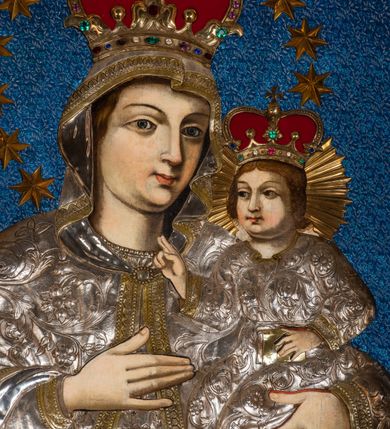 Zdjęcie nr 3: Obraz w kształcie stojącego prostokąta, ujęty rzeźbioną ramą z motywem wici akantowej, przedstawia Madonnę z Dzieciątkiem. Ukazana w półpostaci Maria zwrócona jest na wprost. W trzech czwartych skierowaną w lewo głowę charakteryzuje owalna twarz o wyrazistych rysach z ciemnymi łukami brwi, dużymi, migdałowatymi oczami o wyraźnych, dolnych powiekach, długim, prostym nosie, kształtnymi ustami w subtelnym uśmiechu i wyraźną linią podbródka. Partie szat pokryte są metalowymi osłonami. Maria ma na sobie srebrną, przewiązaną w pasie suknię zdobioną motywem tulipanów, pod szyją krótkie korale. Głowę, ramiona i ręce osłania srebrny, spięty pod szyją owalną broszą płaszcz obszyty złoconymi galonami, zdobiony motywem ukwieconej wici roślinnej. Matka Boska prawą, uniesioną na wysokości piersi ręką o wyprostowanej dłoni i złączonych trzech palcach wskazuje na trzymane na lewej ręce Dzieciątko. Zwrócony w trzech czwartych w prawo Jezus ukazany jest w pozycji siedzącej. Zaokrągloną twarz charakteryzują pełne, wyraziste rysy o dużych oczach z wydanie podkreślonymi liniami powiek, długim nosie, kształtnych ustach w delikatnym uśmiechu, zaróżowionych policzkach oraz wyraźniej linii podbródka. Jezus ma na sobie długą, srebrną, przewiązaną w pasie, zdobioną ukwieconą wicią roślinną suknię o złoconym obszyciu dekoltu i dekoracyjnych galonach przy mankietach dolnej krawędzi. Lewą ręką podtrzymuje małą, zamkniętą, złoconą księgę, prawą unosi w geście błogosławieństwa. Zwieńczone zamkniętymi, kameryzowanymi koronami głowy okalają – promienisty nimb Jezusa, wieniec z 12 gwiazd Marii. Tło niebieskie, z drobnym deseniem. 
