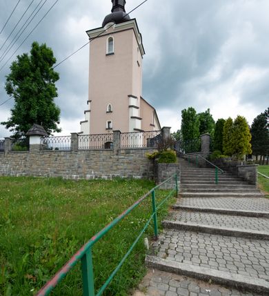 Zdjęcie nr 7: Kościół orientowany, jest budowlą murowaną z cegły i kamienia, na rzucie podłużnym. Korpus nawowy jest jednonawowy i czteroprzęsłowy. Do dwuprzęsłowego, zamkniętego trójbocznie prezbiterium od północy przylega zakrystia. Na osi kościoła, w przedłużeniu północnego przęsła jest czworoboczna, wysunięta przed lico fasady wieża. Od południa do drugiego przęsła korpusu przylega kruchta. Nawa przekryta jest sklepieniem kolebkowym z lunetami wspartymi na pilastrach, w prezbiterium sklepienie krzyżowo-żebrowe z gurtą wspartą na pilastrach. Przestrzeń prezbiterium od korpusu nawowego oddziela półkolista arkada łuku tęczowego i drewniana balustrada. Artykulację ścian na zewnątrz kościoła stanowi zespół dwuuskokowych przypór, w narożach wieży są czterouskokowe, osadzonych na niewysokim cokole. Do wnętrza prowadzą 3 otwory drzwiowe. Dwuskrzydłowe drzwi w fasadzie ujęte są kamiennym obramieniem zamkniętym łukiem koszowym z kluczem pośrodku, wspartym na gzymsie oraz 2 w północnej ścianie kaplicy bocznej. 2 prostokątne prowadzą przez kruchtę i zakrystię. Ściany korpusu nawowego i prezbiterium przeprute są prostokątnymi oknami zamkniętymi łukiem odcinkowym. Okno na zakończeniu prezbiterium jest formie półkoliście zamkniętej niszy z figurą Matki Boskiej. Kształt okien korpusu i prezbiterium został powtórzony w przyległości prezbiterium. Powyżej wejścia na osi wieży małe okienko zamknięte łukiem koszowym, wyżej nisza zamknięta ostrołukowo. W górnej partii wieży znajdują się cztery, duże, zamknięte półkoliście otwory okienne ujęte prostymi obramieniami. Szczyt wierzy obiega gzyms wieńczący z półkolistym łukiem na osi każdej ze ścian. Dach w formie hełmu z latarnią. Dach prezbiterium i korpusu dwuspadowy, kryty blachą, nad pierwszym przęsłem wieżyczka na sygnaturkę. Kruchta z dachem dwuspadowym, przyległość pulpitowym. Tynki w kolorze beżowym i białym. 