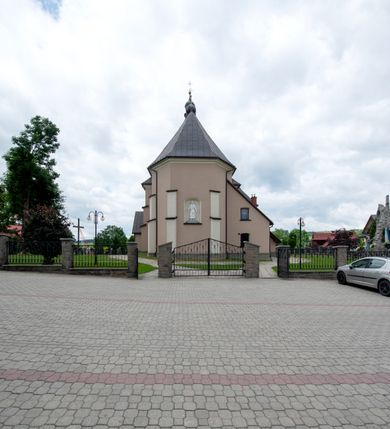 Zdjęcie nr 14: Kościół orientowany, jest budowlą murowaną z cegły i kamienia, na rzucie podłużnym. Korpus nawowy jest jednonawowy i czteroprzęsłowy. Do dwuprzęsłowego, zamkniętego trójbocznie prezbiterium od północy przylega zakrystia. Na osi kościoła, w przedłużeniu północnego przęsła jest czworoboczna, wysunięta przed lico fasady wieża. Od południa do drugiego przęsła korpusu przylega kruchta. Nawa przekryta jest sklepieniem kolebkowym z lunetami wspartymi na pilastrach, w prezbiterium sklepienie krzyżowo-żebrowe z gurtą wspartą na pilastrach. Przestrzeń prezbiterium od korpusu nawowego oddziela półkolista arkada łuku tęczowego i drewniana balustrada. Artykulację ścian na zewnątrz kościoła stanowi zespół dwuuskokowych przypór, w narożach wieży są czterouskokowe, osadzonych na niewysokim cokole. Do wnętrza prowadzą 3 otwory drzwiowe. Dwuskrzydłowe drzwi w fasadzie ujęte są kamiennym obramieniem zamkniętym łukiem koszowym z kluczem pośrodku, wspartym na gzymsie oraz 2 w północnej ścianie kaplicy bocznej. 2 prostokątne prowadzą przez kruchtę i zakrystię. Ściany korpusu nawowego i prezbiterium przeprute są prostokątnymi oknami zamkniętymi łukiem odcinkowym. Okno na zakończeniu prezbiterium jest formie półkoliście zamkniętej niszy z figurą Matki Boskiej. Kształt okien korpusu i prezbiterium został powtórzony w przyległości prezbiterium. Powyżej wejścia na osi wieży małe okienko zamknięte łukiem koszowym, wyżej nisza zamknięta ostrołukowo. W górnej partii wieży znajdują się cztery, duże, zamknięte półkoliście otwory okienne ujęte prostymi obramieniami. Szczyt wierzy obiega gzyms wieńczący z półkolistym łukiem na osi każdej ze ścian. Dach w formie hełmu z latarnią. Dach prezbiterium i korpusu dwuspadowy, kryty blachą, nad pierwszym przęsłem wieżyczka na sygnaturkę. Kruchta z dachem dwuspadowym, przyległość pulpitowym. Tynki w kolorze beżowym i białym. 