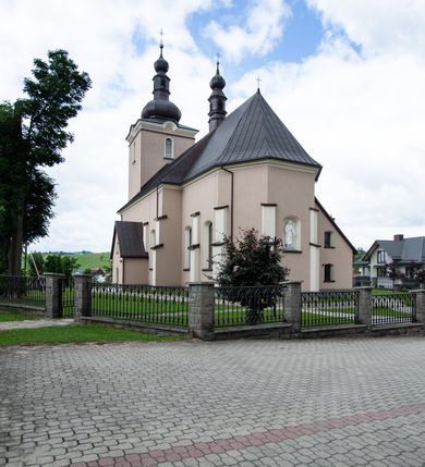 Zdjęcie nr 13: Kościół orientowany, jest budowlą murowaną z cegły i kamienia, na rzucie podłużnym. Korpus nawowy jest jednonawowy i czteroprzęsłowy. Do dwuprzęsłowego, zamkniętego trójbocznie prezbiterium od północy przylega zakrystia. Na osi kościoła, w przedłużeniu północnego przęsła jest czworoboczna, wysunięta przed lico fasady wieża. Od południa do drugiego przęsła korpusu przylega kruchta. Nawa przekryta jest sklepieniem kolebkowym z lunetami wspartymi na pilastrach, w prezbiterium sklepienie krzyżowo-żebrowe z gurtą wspartą na pilastrach. Przestrzeń prezbiterium od korpusu nawowego oddziela półkolista arkada łuku tęczowego i drewniana balustrada. Artykulację ścian na zewnątrz kościoła stanowi zespół dwuuskokowych przypór, w narożach wieży są czterouskokowe, osadzonych na niewysokim cokole. Do wnętrza prowadzą 3 otwory drzwiowe. Dwuskrzydłowe drzwi w fasadzie ujęte są kamiennym obramieniem zamkniętym łukiem koszowym z kluczem pośrodku, wspartym na gzymsie oraz 2 w północnej ścianie kaplicy bocznej. 2 prostokątne prowadzą przez kruchtę i zakrystię. Ściany korpusu nawowego i prezbiterium przeprute są prostokątnymi oknami zamkniętymi łukiem odcinkowym. Okno na zakończeniu prezbiterium jest formie półkoliście zamkniętej niszy z figurą Matki Boskiej. Kształt okien korpusu i prezbiterium został powtórzony w przyległości prezbiterium. Powyżej wejścia na osi wieży małe okienko zamknięte łukiem koszowym, wyżej nisza zamknięta ostrołukowo. W górnej partii wieży znajdują się cztery, duże, zamknięte półkoliście otwory okienne ujęte prostymi obramieniami. Szczyt wierzy obiega gzyms wieńczący z półkolistym łukiem na osi każdej ze ścian. Dach w formie hełmu z latarnią. Dach prezbiterium i korpusu dwuspadowy, kryty blachą, nad pierwszym przęsłem wieżyczka na sygnaturkę. Kruchta z dachem dwuspadowym, przyległość pulpitowym. Tynki w kolorze beżowym i białym. 