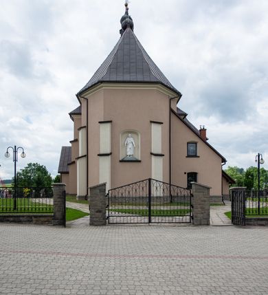 Zdjęcie nr 15: Kościół orientowany, jest budowlą murowaną z cegły i kamienia, na rzucie podłużnym. Korpus nawowy jest jednonawowy i czteroprzęsłowy. Do dwuprzęsłowego, zamkniętego trójbocznie prezbiterium od północy przylega zakrystia. Na osi kościoła, w przedłużeniu północnego przęsła jest czworoboczna, wysunięta przed lico fasady wieża. Od południa do drugiego przęsła korpusu przylega kruchta. Nawa przekryta jest sklepieniem kolebkowym z lunetami wspartymi na pilastrach, w prezbiterium sklepienie krzyżowo-żebrowe z gurtą wspartą na pilastrach. Przestrzeń prezbiterium od korpusu nawowego oddziela półkolista arkada łuku tęczowego i drewniana balustrada. Artykulację ścian na zewnątrz kościoła stanowi zespół dwuuskokowych przypór, w narożach wieży są czterouskokowe, osadzonych na niewysokim cokole. Do wnętrza prowadzą 3 otwory drzwiowe. Dwuskrzydłowe drzwi w fasadzie ujęte są kamiennym obramieniem zamkniętym łukiem koszowym z kluczem pośrodku, wspartym na gzymsie oraz 2 w północnej ścianie kaplicy bocznej. 2 prostokątne prowadzą przez kruchtę i zakrystię. Ściany korpusu nawowego i prezbiterium przeprute są prostokątnymi oknami zamkniętymi łukiem odcinkowym. Okno na zakończeniu prezbiterium jest formie półkoliście zamkniętej niszy z figurą Matki Boskiej. Kształt okien korpusu i prezbiterium został powtórzony w przyległości prezbiterium. Powyżej wejścia na osi wieży małe okienko zamknięte łukiem koszowym, wyżej nisza zamknięta ostrołukowo. W górnej partii wieży znajdują się cztery, duże, zamknięte półkoliście otwory okienne ujęte prostymi obramieniami. Szczyt wierzy obiega gzyms wieńczący z półkolistym łukiem na osi każdej ze ścian. Dach w formie hełmu z latarnią. Dach prezbiterium i korpusu dwuspadowy, kryty blachą, nad pierwszym przęsłem wieżyczka na sygnaturkę. Kruchta z dachem dwuspadowym, przyległość pulpitowym. Tynki w kolorze beżowym i białym. 