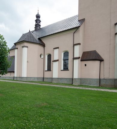 Zdjęcie nr 12: Kościół orientowany, jest budowlą murowaną z cegły i kamienia, na rzucie podłużnym. Korpus nawowy jest jednonawowy i czteroprzęsłowy. Do dwuprzęsłowego, zamkniętego trójbocznie prezbiterium od północy przylega zakrystia. Na osi kościoła, w przedłużeniu północnego przęsła jest czworoboczna, wysunięta przed lico fasady wieża. Od południa do drugiego przęsła korpusu przylega kruchta. Nawa przekryta jest sklepieniem kolebkowym z lunetami wspartymi na pilastrach, w prezbiterium sklepienie krzyżowo-żebrowe z gurtą wspartą na pilastrach. Przestrzeń prezbiterium od korpusu nawowego oddziela półkolista arkada łuku tęczowego i drewniana balustrada. Artykulację ścian na zewnątrz kościoła stanowi zespół dwuuskokowych przypór, w narożach wieży są czterouskokowe, osadzonych na niewysokim cokole. Do wnętrza prowadzą 3 otwory drzwiowe. Dwuskrzydłowe drzwi w fasadzie ujęte są kamiennym obramieniem zamkniętym łukiem koszowym z kluczem pośrodku, wspartym na gzymsie oraz 2 w północnej ścianie kaplicy bocznej. 2 prostokątne prowadzą przez kruchtę i zakrystię. Ściany korpusu nawowego i prezbiterium przeprute są prostokątnymi oknami zamkniętymi łukiem odcinkowym. Okno na zakończeniu prezbiterium jest formie półkoliście zamkniętej niszy z figurą Matki Boskiej. Kształt okien korpusu i prezbiterium został powtórzony w przyległości prezbiterium. Powyżej wejścia na osi wieży małe okienko zamknięte łukiem koszowym, wyżej nisza zamknięta ostrołukowo. W górnej partii wieży znajdują się cztery, duże, zamknięte półkoliście otwory okienne ujęte prostymi obramieniami. Szczyt wierzy obiega gzyms wieńczący z półkolistym łukiem na osi każdej ze ścian. Dach w formie hełmu z latarnią. Dach prezbiterium i korpusu dwuspadowy, kryty blachą, nad pierwszym przęsłem wieżyczka na sygnaturkę. Kruchta z dachem dwuspadowym, przyległość pulpitowym. Tynki w kolorze beżowym i białym. 