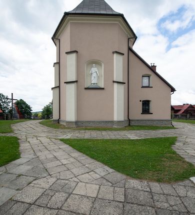 Zdjęcie nr 16: Kościół orientowany, jest budowlą murowaną z cegły i kamienia, na rzucie podłużnym. Korpus nawowy jest jednonawowy i czteroprzęsłowy. Do dwuprzęsłowego, zamkniętego trójbocznie prezbiterium od północy przylega zakrystia. Na osi kościoła, w przedłużeniu północnego przęsła jest czworoboczna, wysunięta przed lico fasady wieża. Od południa do drugiego przęsła korpusu przylega kruchta. Nawa przekryta jest sklepieniem kolebkowym z lunetami wspartymi na pilastrach, w prezbiterium sklepienie krzyżowo-żebrowe z gurtą wspartą na pilastrach. Przestrzeń prezbiterium od korpusu nawowego oddziela półkolista arkada łuku tęczowego i drewniana balustrada. Artykulację ścian na zewnątrz kościoła stanowi zespół dwuuskokowych przypór, w narożach wieży są czterouskokowe, osadzonych na niewysokim cokole. Do wnętrza prowadzą 3 otwory drzwiowe. Dwuskrzydłowe drzwi w fasadzie ujęte są kamiennym obramieniem zamkniętym łukiem koszowym z kluczem pośrodku, wspartym na gzymsie oraz 2 w północnej ścianie kaplicy bocznej. 2 prostokątne prowadzą przez kruchtę i zakrystię. Ściany korpusu nawowego i prezbiterium przeprute są prostokątnymi oknami zamkniętymi łukiem odcinkowym. Okno na zakończeniu prezbiterium jest formie półkoliście zamkniętej niszy z figurą Matki Boskiej. Kształt okien korpusu i prezbiterium został powtórzony w przyległości prezbiterium. Powyżej wejścia na osi wieży małe okienko zamknięte łukiem koszowym, wyżej nisza zamknięta ostrołukowo. W górnej partii wieży znajdują się cztery, duże, zamknięte półkoliście otwory okienne ujęte prostymi obramieniami. Szczyt wierzy obiega gzyms wieńczący z półkolistym łukiem na osi każdej ze ścian. Dach w formie hełmu z latarnią. Dach prezbiterium i korpusu dwuspadowy, kryty blachą, nad pierwszym przęsłem wieżyczka na sygnaturkę. Kruchta z dachem dwuspadowym, przyległość pulpitowym. Tynki w kolorze beżowym i białym. 