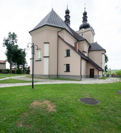 Zdjęcie nr 50: Kościół orientowany, jest budowlą murowaną z cegły i kamienia, na rzucie podłużnym. Korpus nawowy jest jednonawowy i czteroprzęsłowy. Do dwuprzęsłowego, zamkniętego trójbocznie prezbiterium od północy przylega zakrystia. Na osi kościoła, w przedłużeniu północnego przęsła jest czworoboczna, wysunięta przed lico fasady wieża. Od południa do drugiego przęsła korpusu przylega kruchta. Nawa przekryta jest sklepieniem kolebkowym z lunetami wspartymi na pilastrach, w prezbiterium sklepienie krzyżowo-żebrowe z gurtą wspartą na pilastrach. Przestrzeń prezbiterium od korpusu nawowego oddziela półkolista arkada łuku tęczowego i drewniana balustrada. Artykulację ścian na zewnątrz kościoła stanowi zespół dwuuskokowych przypór, w narożach wieży są czterouskokowe, osadzonych na niewysokim cokole. Do wnętrza prowadzą 3 otwory drzwiowe. Dwuskrzydłowe drzwi w fasadzie ujęte są kamiennym obramieniem zamkniętym łukiem koszowym z kluczem pośrodku, wspartym na gzymsie oraz 2 w północnej ścianie kaplicy bocznej. 2 prostokątne prowadzą przez kruchtę i zakrystię. Ściany korpusu nawowego i prezbiterium przeprute są prostokątnymi oknami zamkniętymi łukiem odcinkowym. Okno na zakończeniu prezbiterium jest formie półkoliście zamkniętej niszy z figurą Matki Boskiej. Kształt okien korpusu i prezbiterium został powtórzony w przyległości prezbiterium. Powyżej wejścia na osi wieży małe okienko zamknięte łukiem koszowym, wyżej nisza zamknięta ostrołukowo. W górnej partii wieży znajdują się cztery, duże, zamknięte półkoliście otwory okienne ujęte prostymi obramieniami. Szczyt wierzy obiega gzyms wieńczący z półkolistym łukiem na osi każdej ze ścian. Dach w formie hełmu z latarnią. Dach prezbiterium i korpusu dwuspadowy, kryty blachą, nad pierwszym przęsłem wieżyczka na sygnaturkę. Kruchta z dachem dwuspadowym, przyległość pulpitowym. Tynki w kolorze beżowym i białym. 