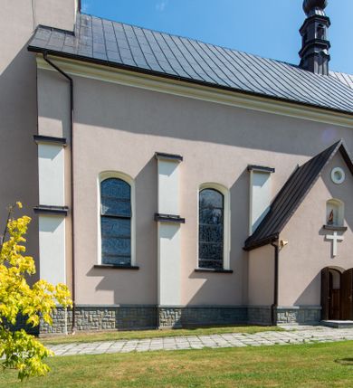 Zdjęcie nr 19: Kościół orientowany, jest budowlą murowaną z cegły i kamienia, na rzucie podłużnym. Korpus nawowy jest jednonawowy i czteroprzęsłowy. Do dwuprzęsłowego, zamkniętego trójbocznie prezbiterium od północy przylega zakrystia. Na osi kościoła, w przedłużeniu północnego przęsła jest czworoboczna, wysunięta przed lico fasady wieża. Od południa do drugiego przęsła korpusu przylega kruchta. Nawa przekryta jest sklepieniem kolebkowym z lunetami wspartymi na pilastrach, w prezbiterium sklepienie krzyżowo-żebrowe z gurtą wspartą na pilastrach. Przestrzeń prezbiterium od korpusu nawowego oddziela półkolista arkada łuku tęczowego i drewniana balustrada. Artykulację ścian na zewnątrz kościoła stanowi zespół dwuuskokowych przypór, w narożach wieży są czterouskokowe, osadzonych na niewysokim cokole. Do wnętrza prowadzą 3 otwory drzwiowe. Dwuskrzydłowe drzwi w fasadzie ujęte są kamiennym obramieniem zamkniętym łukiem koszowym z kluczem pośrodku, wspartym na gzymsie oraz 2 w północnej ścianie kaplicy bocznej. 2 prostokątne prowadzą przez kruchtę i zakrystię. Ściany korpusu nawowego i prezbiterium przeprute są prostokątnymi oknami zamkniętymi łukiem odcinkowym. Okno na zakończeniu prezbiterium jest formie półkoliście zamkniętej niszy z figurą Matki Boskiej. Kształt okien korpusu i prezbiterium został powtórzony w przyległości prezbiterium. Powyżej wejścia na osi wieży małe okienko zamknięte łukiem koszowym, wyżej nisza zamknięta ostrołukowo. W górnej partii wieży znajdują się cztery, duże, zamknięte półkoliście otwory okienne ujęte prostymi obramieniami. Szczyt wierzy obiega gzyms wieńczący z półkolistym łukiem na osi każdej ze ścian. Dach w formie hełmu z latarnią. Dach prezbiterium i korpusu dwuspadowy, kryty blachą, nad pierwszym przęsłem wieżyczka na sygnaturkę. Kruchta z dachem dwuspadowym, przyległość pulpitowym. Tynki w kolorze beżowym i białym. 