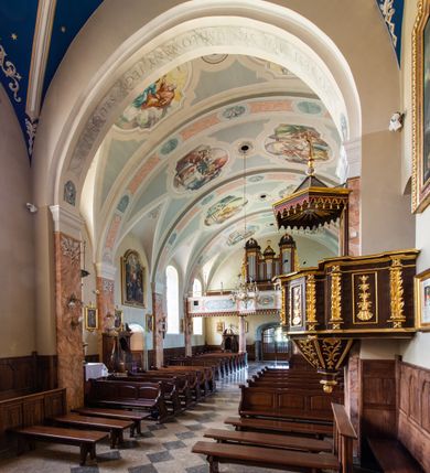 Zdjęcie nr 44: Kościół orientowany, jest budowlą murowaną z cegły i kamienia, na rzucie podłużnym. Korpus nawowy jest jednonawowy i czteroprzęsłowy. Do dwuprzęsłowego, zamkniętego trójbocznie prezbiterium od północy przylega zakrystia. Na osi kościoła, w przedłużeniu północnego przęsła jest czworoboczna, wysunięta przed lico fasady wieża. Od południa do drugiego przęsła korpusu przylega kruchta. Nawa przekryta jest sklepieniem kolebkowym z lunetami wspartymi na pilastrach, w prezbiterium sklepienie krzyżowo-żebrowe z gurtą wspartą na pilastrach. Przestrzeń prezbiterium od korpusu nawowego oddziela półkolista arkada łuku tęczowego i drewniana balustrada. Artykulację ścian na zewnątrz kościoła stanowi zespół dwuuskokowych przypór, w narożach wieży są czterouskokowe, osadzonych na niewysokim cokole. Do wnętrza prowadzą 3 otwory drzwiowe. Dwuskrzydłowe drzwi w fasadzie ujęte są kamiennym obramieniem zamkniętym łukiem koszowym z kluczem pośrodku, wspartym na gzymsie oraz 2 w północnej ścianie kaplicy bocznej. 2 prostokątne prowadzą przez kruchtę i zakrystię. Ściany korpusu nawowego i prezbiterium przeprute są prostokątnymi oknami zamkniętymi łukiem odcinkowym. Okno na zakończeniu prezbiterium jest formie półkoliście zamkniętej niszy z figurą Matki Boskiej. Kształt okien korpusu i prezbiterium został powtórzony w przyległości prezbiterium. Powyżej wejścia na osi wieży małe okienko zamknięte łukiem koszowym, wyżej nisza zamknięta ostrołukowo. W górnej partii wieży znajdują się cztery, duże, zamknięte półkoliście otwory okienne ujęte prostymi obramieniami. Szczyt wierzy obiega gzyms wieńczący z półkolistym łukiem na osi każdej ze ścian. Dach w formie hełmu z latarnią. Dach prezbiterium i korpusu dwuspadowy, kryty blachą, nad pierwszym przęsłem wieżyczka na sygnaturkę. Kruchta z dachem dwuspadowym, przyległość pulpitowym. Tynki w kolorze beżowym i białym. 