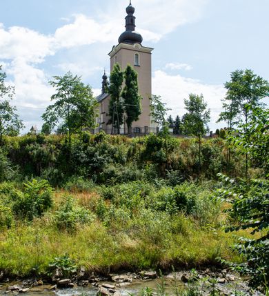 Zdjęcie nr 4: Kościół orientowany, jest budowlą murowaną z cegły i kamienia, na rzucie podłużnym. Korpus nawowy jest jednonawowy i czteroprzęsłowy. Do dwuprzęsłowego, zamkniętego trójbocznie prezbiterium od północy przylega zakrystia. Na osi kościoła, w przedłużeniu północnego przęsła jest czworoboczna, wysunięta przed lico fasady wieża. Od południa do drugiego przęsła korpusu przylega kruchta. Nawa przekryta jest sklepieniem kolebkowym z lunetami wspartymi na pilastrach, w prezbiterium sklepienie krzyżowo-żebrowe z gurtą wspartą na pilastrach. Przestrzeń prezbiterium od korpusu nawowego oddziela półkolista arkada łuku tęczowego i drewniana balustrada. Artykulację ścian na zewnątrz kościoła stanowi zespół dwuuskokowych przypór, w narożach wieży są czterouskokowe, osadzonych na niewysokim cokole. Do wnętrza prowadzą 3 otwory drzwiowe. Dwuskrzydłowe drzwi w fasadzie ujęte są kamiennym obramieniem zamkniętym łukiem koszowym z kluczem pośrodku, wspartym na gzymsie oraz 2 w północnej ścianie kaplicy bocznej. 2 prostokątne prowadzą przez kruchtę i zakrystię. Ściany korpusu nawowego i prezbiterium przeprute są prostokątnymi oknami zamkniętymi łukiem odcinkowym. Okno na zakończeniu prezbiterium jest formie półkoliście zamkniętej niszy z figurą Matki Boskiej. Kształt okien korpusu i prezbiterium został powtórzony w przyległości prezbiterium. Powyżej wejścia na osi wieży małe okienko zamknięte łukiem koszowym, wyżej nisza zamknięta ostrołukowo. W górnej partii wieży znajdują się cztery, duże, zamknięte półkoliście otwory okienne ujęte prostymi obramieniami. Szczyt wierzy obiega gzyms wieńczący z półkolistym łukiem na osi każdej ze ścian. Dach w formie hełmu z latarnią. Dach prezbiterium i korpusu dwuspadowy, kryty blachą, nad pierwszym przęsłem wieżyczka na sygnaturkę. Kruchta z dachem dwuspadowym, przyległość pulpitowym. Tynki w kolorze beżowym i białym. 