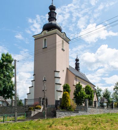 Zdjęcie nr 9: Kościół orientowany, jest budowlą murowaną z cegły i kamienia, na rzucie podłużnym. Korpus nawowy jest jednonawowy i czteroprzęsłowy. Do dwuprzęsłowego, zamkniętego trójbocznie prezbiterium od północy przylega zakrystia. Na osi kościoła, w przedłużeniu północnego przęsła jest czworoboczna, wysunięta przed lico fasady wieża. Od południa do drugiego przęsła korpusu przylega kruchta. Nawa przekryta jest sklepieniem kolebkowym z lunetami wspartymi na pilastrach, w prezbiterium sklepienie krzyżowo-żebrowe z gurtą wspartą na pilastrach. Przestrzeń prezbiterium od korpusu nawowego oddziela półkolista arkada łuku tęczowego i drewniana balustrada. Artykulację ścian na zewnątrz kościoła stanowi zespół dwuuskokowych przypór, w narożach wieży są czterouskokowe, osadzonych na niewysokim cokole. Do wnętrza prowadzą 3 otwory drzwiowe. Dwuskrzydłowe drzwi w fasadzie ujęte są kamiennym obramieniem zamkniętym łukiem koszowym z kluczem pośrodku, wspartym na gzymsie oraz 2 w północnej ścianie kaplicy bocznej. 2 prostokątne prowadzą przez kruchtę i zakrystię. Ściany korpusu nawowego i prezbiterium przeprute są prostokątnymi oknami zamkniętymi łukiem odcinkowym. Okno na zakończeniu prezbiterium jest formie półkoliście zamkniętej niszy z figurą Matki Boskiej. Kształt okien korpusu i prezbiterium został powtórzony w przyległości prezbiterium. Powyżej wejścia na osi wieży małe okienko zamknięte łukiem koszowym, wyżej nisza zamknięta ostrołukowo. W górnej partii wieży znajdują się cztery, duże, zamknięte półkoliście otwory okienne ujęte prostymi obramieniami. Szczyt wierzy obiega gzyms wieńczący z półkolistym łukiem na osi każdej ze ścian. Dach w formie hełmu z latarnią. Dach prezbiterium i korpusu dwuspadowy, kryty blachą, nad pierwszym przęsłem wieżyczka na sygnaturkę. Kruchta z dachem dwuspadowym, przyległość pulpitowym. Tynki w kolorze beżowym i białym. 
