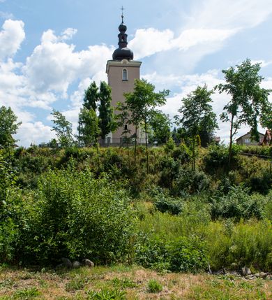 Zdjęcie nr 53: Kościół orientowany, jest budowlą murowaną z cegły i kamienia, na rzucie podłużnym. Korpus nawowy jest jednonawowy i czteroprzęsłowy. Do dwuprzęsłowego, zamkniętego trójbocznie prezbiterium od północy przylega zakrystia. Na osi kościoła, w przedłużeniu północnego przęsła jest czworoboczna, wysunięta przed lico fasady wieża. Od południa do drugiego przęsła korpusu przylega kruchta. Nawa przekryta jest sklepieniem kolebkowym z lunetami wspartymi na pilastrach, w prezbiterium sklepienie krzyżowo-żebrowe z gurtą wspartą na pilastrach. Przestrzeń prezbiterium od korpusu nawowego oddziela półkolista arkada łuku tęczowego i drewniana balustrada. Artykulację ścian na zewnątrz kościoła stanowi zespół dwuuskokowych przypór, w narożach wieży są czterouskokowe, osadzonych na niewysokim cokole. Do wnętrza prowadzą 3 otwory drzwiowe. Dwuskrzydłowe drzwi w fasadzie ujęte są kamiennym obramieniem zamkniętym łukiem koszowym z kluczem pośrodku, wspartym na gzymsie oraz 2 w północnej ścianie kaplicy bocznej. 2 prostokątne prowadzą przez kruchtę i zakrystię. Ściany korpusu nawowego i prezbiterium przeprute są prostokątnymi oknami zamkniętymi łukiem odcinkowym. Okno na zakończeniu prezbiterium jest formie półkoliście zamkniętej niszy z figurą Matki Boskiej. Kształt okien korpusu i prezbiterium został powtórzony w przyległości prezbiterium. Powyżej wejścia na osi wieży małe okienko zamknięte łukiem koszowym, wyżej nisza zamknięta ostrołukowo. W górnej partii wieży znajdują się cztery, duże, zamknięte półkoliście otwory okienne ujęte prostymi obramieniami. Szczyt wierzy obiega gzyms wieńczący z półkolistym łukiem na osi każdej ze ścian. Dach w formie hełmu z latarnią. Dach prezbiterium i korpusu dwuspadowy, kryty blachą, nad pierwszym przęsłem wieżyczka na sygnaturkę. Kruchta z dachem dwuspadowym, przyległość pulpitowym. Tynki w kolorze beżowym i białym. 