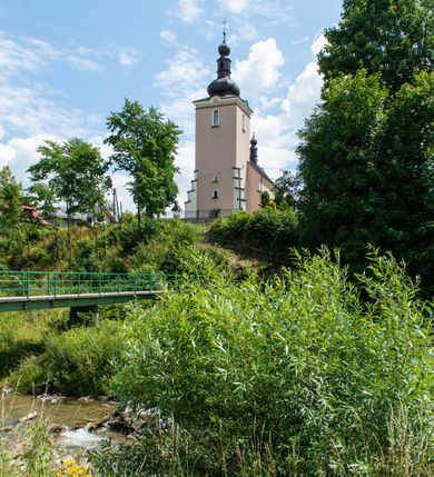 Zdjęcie nr 5: Kościół orientowany, jest budowlą murowaną z cegły i kamienia, na rzucie podłużnym. Korpus nawowy jest jednonawowy i czteroprzęsłowy. Do dwuprzęsłowego, zamkniętego trójbocznie prezbiterium od północy przylega zakrystia. Na osi kościoła, w przedłużeniu północnego przęsła jest czworoboczna, wysunięta przed lico fasady wieża. Od południa do drugiego przęsła korpusu przylega kruchta. Nawa przekryta jest sklepieniem kolebkowym z lunetami wspartymi na pilastrach, w prezbiterium sklepienie krzyżowo-żebrowe z gurtą wspartą na pilastrach. Przestrzeń prezbiterium od korpusu nawowego oddziela półkolista arkada łuku tęczowego i drewniana balustrada. Artykulację ścian na zewnątrz kościoła stanowi zespół dwuuskokowych przypór, w narożach wieży są czterouskokowe, osadzonych na niewysokim cokole. Do wnętrza prowadzą 3 otwory drzwiowe. Dwuskrzydłowe drzwi w fasadzie ujęte są kamiennym obramieniem zamkniętym łukiem koszowym z kluczem pośrodku, wspartym na gzymsie oraz 2 w północnej ścianie kaplicy bocznej. 2 prostokątne prowadzą przez kruchtę i zakrystię. Ściany korpusu nawowego i prezbiterium przeprute są prostokątnymi oknami zamkniętymi łukiem odcinkowym. Okno na zakończeniu prezbiterium jest formie półkoliście zamkniętej niszy z figurą Matki Boskiej. Kształt okien korpusu i prezbiterium został powtórzony w przyległości prezbiterium. Powyżej wejścia na osi wieży małe okienko zamknięte łukiem koszowym, wyżej nisza zamknięta ostrołukowo. W górnej partii wieży znajdują się cztery, duże, zamknięte półkoliście otwory okienne ujęte prostymi obramieniami. Szczyt wierzy obiega gzyms wieńczący z półkolistym łukiem na osi każdej ze ścian. Dach w formie hełmu z latarnią. Dach prezbiterium i korpusu dwuspadowy, kryty blachą, nad pierwszym przęsłem wieżyczka na sygnaturkę. Kruchta z dachem dwuspadowym, przyległość pulpitowym. Tynki w kolorze beżowym i białym. 