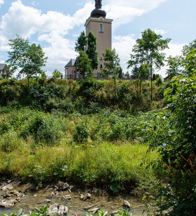 Zdjęcie nr 3: Kościół orientowany, jest budowlą murowaną z cegły i kamienia, na rzucie podłużnym. Korpus nawowy jest jednonawowy i czteroprzęsłowy. Do dwuprzęsłowego, zamkniętego trójbocznie prezbiterium od północy przylega zakrystia. Na osi kościoła, w przedłużeniu północnego przęsła jest czworoboczna, wysunięta przed lico fasady wieża. Od południa do drugiego przęsła korpusu przylega kruchta. Nawa przekryta jest sklepieniem kolebkowym z lunetami wspartymi na pilastrach, w prezbiterium sklepienie krzyżowo-żebrowe z gurtą wspartą na pilastrach. Przestrzeń prezbiterium od korpusu nawowego oddziela półkolista arkada łuku tęczowego i drewniana balustrada. Artykulację ścian na zewnątrz kościoła stanowi zespół dwuuskokowych przypór, w narożach wieży są czterouskokowe, osadzonych na niewysokim cokole. Do wnętrza prowadzą 3 otwory drzwiowe. Dwuskrzydłowe drzwi w fasadzie ujęte są kamiennym obramieniem zamkniętym łukiem koszowym z kluczem pośrodku, wspartym na gzymsie oraz 2 w północnej ścianie kaplicy bocznej. 2 prostokątne prowadzą przez kruchtę i zakrystię. Ściany korpusu nawowego i prezbiterium przeprute są prostokątnymi oknami zamkniętymi łukiem odcinkowym. Okno na zakończeniu prezbiterium jest formie półkoliście zamkniętej niszy z figurą Matki Boskiej. Kształt okien korpusu i prezbiterium został powtórzony w przyległości prezbiterium. Powyżej wejścia na osi wieży małe okienko zamknięte łukiem koszowym, wyżej nisza zamknięta ostrołukowo. W górnej partii wieży znajdują się cztery, duże, zamknięte półkoliście otwory okienne ujęte prostymi obramieniami. Szczyt wierzy obiega gzyms wieńczący z półkolistym łukiem na osi każdej ze ścian. Dach w formie hełmu z latarnią. Dach prezbiterium i korpusu dwuspadowy, kryty blachą, nad pierwszym przęsłem wieżyczka na sygnaturkę. Kruchta z dachem dwuspadowym, przyległość pulpitowym. Tynki w kolorze beżowym i białym. 