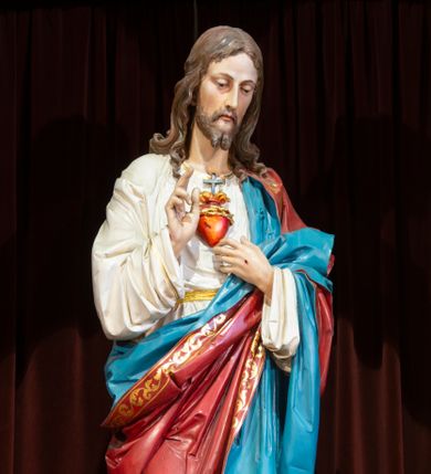 Zdjęcie nr 1: Całopostaciowa, pełnoplastyczna, ustawiona na niewysokiej, prostopadłościennej podstawie  figura Najświętszego Serca Jezusa. Jezus, stojąc w kontrapoście, zwrócony jest na wprost. Lekko przechyloną w lewo głowę charakteryzuje pociągła twarz o delikatnych rysach i krótkim zaroście. Długie, falowane włosy wspierają się na ramionach. Jezus ubrany jest w białą suknię przewiązaną żółtym paskiem i przewieszony przez lewe ramię, osłaniający biodra i nogi czerwony płaszcz o złoconym galonie i niebieskiej podszewce. Prawą rękę unosi w geście błogosławieństwa, lewą wskazuje na gorejące na piersiach serce. Na dłoniach widoczne są ślady ran.    