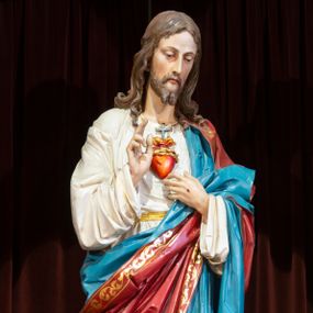Zdjęcie nr 1: Całopostaciowa, pełnoplastyczna, ustawiona na niewysokiej, prostopadłościennej podstawie  figura Najświętszego Serca Jezusa. Jezus, stojąc w kontrapoście, zwrócony jest na wprost. Lekko przechyloną w lewo głowę charakteryzuje pociągła twarz o delikatnych rysach i krótkim zaroście. Długie, falowane włosy wspierają się na ramionach. Jezus ubrany jest w białą suknię przewiązaną żółtym paskiem i przewieszony przez lewe ramię, osłaniający biodra i nogi czerwony płaszcz o złoconym galonie i niebieskiej podszewce. Prawą rękę unosi w geście błogosławieństwa, lewą wskazuje na gorejące na piersiach serce. Na dłoniach widoczne są ślady ran.    