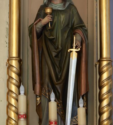 Zdjęcie nr 1: Rzeźba św. Barbary, pełnoplastyczna, ustawiona na niskiej, prostopadłościennej podstawie o ściętych narożach. Święta ustawiona jest frontalnie, w delikatnym kontrapoście z prawą nogą ugiętą w kolanie, z głową pochyloną w dół. W prawej ręce trzyma przed sobą kielich, a w lewej miecz zwrócony ostrzem do dołu. Twarz pociągła o wydatnym podbródku, długi nos, duże oczy, rysy delikatnie rzeźbione. Włosy ciemnobrązowe, długie, falowane, opadające na plecy i barki. Święta ubrana jest w szarą suknię z długimi rękawami i ciemnozielony płaszcz z bordową podszewką, zakładany przez głowę. Płaszcz zdobiony jest na brzegu szeroką bordiurą w formie stylizowanej wici roślinnej. Na głowie ma założoną jasnoszarą chustę oraz złotą, otwartą koronę. Na stopach ma ciemnobrązowe buty. Polichromia naturalistyczna w odsłoniętych partiach ciała. 
