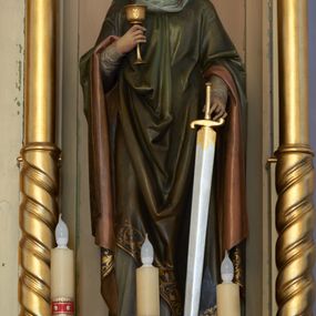 Zdjęcie nr 1: Rzeźba św. Barbary, pełnoplastyczna, ustawiona na niskiej, prostopadłościennej podstawie o ściętych narożach. Święta ustawiona jest frontalnie, w delikatnym kontrapoście z prawą nogą ugiętą w kolanie, z głową pochyloną w dół. W prawej ręce trzyma przed sobą kielich, a w lewej miecz zwrócony ostrzem do dołu. Twarz pociągła o wydatnym podbródku, długi nos, duże oczy, rysy delikatnie rzeźbione. Włosy ciemnobrązowe, długie, falowane, opadające na plecy i barki. Święta ubrana jest w szarą suknię z długimi rękawami i ciemnozielony płaszcz z bordową podszewką, zakładany przez głowę. Płaszcz zdobiony jest na brzegu szeroką bordiurą w formie stylizowanej wici roślinnej. Na głowie ma założoną jasnoszarą chustę oraz złotą, otwartą koronę. Na stopach ma ciemnobrązowe buty. Polichromia naturalistyczna w odsłoniętych partiach ciała. 
