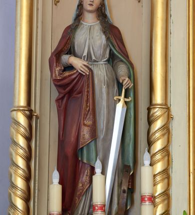 Zdjęcie nr 1: Rzeźba św. Katarzyny Aleksandryjskiej, pełnoplastyczna, ustawiona na niskiej, prostopadłościennej podstawie o ściętych narożach. Święta ustawiona jest frontalnie, w delikatnym  kontrapoście, z głową przechyloną w stronę lewego ramienia. W lewej dłoni trzyma miecz zwrócony ostrzem do dołu, a prawą ugiętą w łokciu przytrzymuje brzeg płaszcza. Twarz owalna o wydatnym podbródku, długi nos, duże oczy, rysy delikatnie rzeźbione. Włosy ciemnobrązowe, długie, falowane, opadające na plecy i ramiona. Święta ubrana jest w szarą suknię z długimi rękawami, przewiązaną w talii i bordowy płaszcz z zieloną podszewką. Płaszcz zdobiony jest na brzegu szeroką bordiurą w formie stylizowanej wici roślinnej. Na głowie święta ma założony jasnoszary welon oraz złotą, otwartą koronę, a na stopach ciemnobrązowe sandały. W podstawie, po lewej stronie świętej zatknięte jest koło. Polichromia naturalistyczna w odsłoniętych partiach ciała. 
