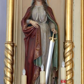 Zdjęcie nr 1: Rzeźba św. Katarzyny Aleksandryjskiej, pełnoplastyczna, ustawiona na niskiej, prostopadłościennej podstawie o ściętych narożach. Święta ustawiona jest frontalnie, w delikatnym  kontrapoście, z głową przechyloną w stronę lewego ramienia. W lewej dłoni trzyma miecz zwrócony ostrzem do dołu, a prawą ugiętą w łokciu przytrzymuje brzeg płaszcza. Twarz owalna o wydatnym podbródku, długi nos, duże oczy, rysy delikatnie rzeźbione. Włosy ciemnobrązowe, długie, falowane, opadające na plecy i ramiona. Święta ubrana jest w szarą suknię z długimi rękawami, przewiązaną w talii i bordowy płaszcz z zieloną podszewką. Płaszcz zdobiony jest na brzegu szeroką bordiurą w formie stylizowanej wici roślinnej. Na głowie święta ma założony jasnoszary welon oraz złotą, otwartą koronę, a na stopach ciemnobrązowe sandały. W podstawie, po lewej stronie świętej zatknięte jest koło. Polichromia naturalistyczna w odsłoniętych partiach ciała. 
