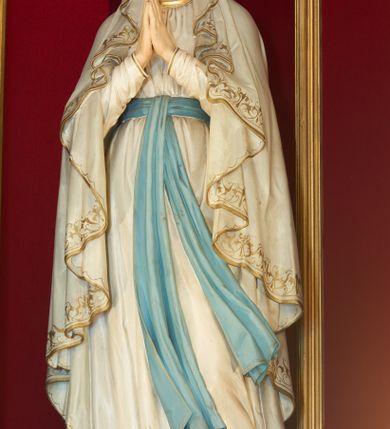 Zdjęcie nr 1: Rzeźba pełnoplastyczna, ustawiona na niskim postumencie z przedstawieniem Matki Boskiej z Lourdes. Maria ukazana frontalnie, w postawie stojącej, w delikatnym kontrapoście, z lewą nogą ugiętą w kolanie i z rękami złożonymi w geście modlitwy. Matka Boska ubrana jest w białą suknię ze złotą lamówką u szyi, przewiązaną w talii błękitną szarfą oraz biały płaszcz założony na głowę i spływający delikatnymi falami wzdłuż całej postaci, na brzegach obwiedziony złotą i dekoracyjną bordiurą. Twarz owalna z długim i wąskim nosem, z wyraźnie zaznaczonym podbródkiem oraz dużymi i niebieskimi oczami; na głowie korona w typie otwartym. Polichromia w odsłoniętych partiach ciała naturalistyczna.


