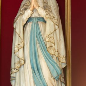 Zdjęcie nr 1: Rzeźba pełnoplastyczna, ustawiona na niskim postumencie z przedstawieniem Matki Boskiej z Lourdes. Maria ukazana frontalnie, w postawie stojącej, w delikatnym kontrapoście, z lewą nogą ugiętą w kolanie i z rękami złożonymi w geście modlitwy. Matka Boska ubrana jest w białą suknię ze złotą lamówką u szyi, przewiązaną w talii błękitną szarfą oraz biały płaszcz założony na głowę i spływający delikatnymi falami wzdłuż całej postaci, na brzegach obwiedziony złotą i dekoracyjną bordiurą. Twarz owalna z długim i wąskim nosem, z wyraźnie zaznaczonym podbródkiem oraz dużymi i niebieskimi oczami; na głowie korona w typie otwartym. Polichromia w odsłoniętych partiach ciała naturalistyczna.


