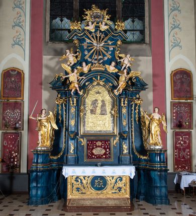 Zdjęcie nr 2: Nastawa architektoniczna, jednoosiowa, jednokondygnacyjna ze zwieńczeniem, ustawiona na wysokim cokole. Ołtarz drewniany, prostopadłościenny, o falistych bokach, mensa drewniana z marmurowym portatylem w drewnianej obudowie. Antepedium ujęte szeroką ramą dekorowaną płaskorzeźbionym motywem wieńca przepasanego wstęgą, w polu medalion z monogramem Marii ujęty wychodzącymi spod jego spodu wiązankami utworzonymi z suchego, wijącego się akantu z karbowaną wstęgą. Nastawa ujęta dwiema parami pilastrów, wewnętrzną z wolutowymi wspornikami w połowie wysokości trzonów oraz zewnętrzną parą o trzonach w formie spływów wolutowych przechodzących w silnie wysunięte do przodu partie cokołu, na których znajdują się postumenty dla figur aniołów. Podpory dźwigają niepełne belkowanie wyłamane na ich osi, nad polem środkowym wyłącznie gzyms przyjmujący kształt łuku półkolistego. Na belkowaniu fragmenty przerwanego, falistego przyczółka oraz para aniołów z kartuszami. W polu środkowym wnęka w formie stojącego prostokąta zamkniętego łukiem segmentowym nadwieszonym z uskokiem, w profilowanej ramie z mniejszym obrazem Matki Boskiej z Dzieciątkiem w kształcie stojącego prostokąta. Poniżej gablota na wota w formie leżącego prostokąta ujęta profilowaną ramą. Kondygnacja flankowana uszami utworzonymi z rocaille'u. Zwieńczenie w formie zbliżonej do kwadratu o wklęsłych bokach przechodzących w woluty, na których siedzą putta, zamknięte fragmentem falistego gzymsu wyłamanego po bokach, na nim rokokowy kartusz z monogramem IHS w polu, flankowany rocaille'em. W polu zwieńczenia rzeźba gołębicy Ducha Świętego w glorii promienistej i otoku z obłoków, w towarzystwie czterech uskrzydlonych główek anielskich. Wszystkie pilastry oraz łuki dekorowane elementami rocaille'u. Struktura w całości polichromowana na sposób imitujący marmoryzację w kolorze niebieskim, ornamentyka i profile złocone, detal srebrzony. 
Wewnątrz skrzyni ołtarzowej napisy „MALOWAŁ LUDWIK WÓJCIK Z KRAKOWA 2 / XI 1914”; „LUDWIK WÓJCIK”; „MALOWALI STOPY”; „23.02.1994 / KONSERWOWALI / KAZIMIERZ PATEJUK / EWA I JERZY SUCHWAŁKO / Z WARSZAWY / PROBOSZCZOWAŁ / KS. BOLESŁAW WAWAK”. 