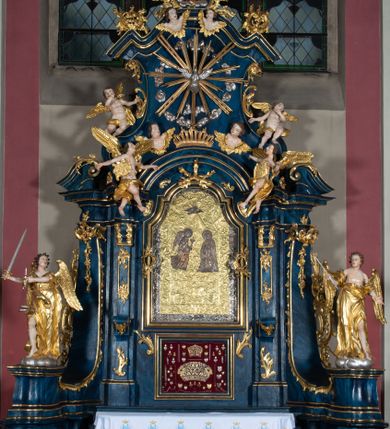 Zdjęcie nr 1: Nastawa architektoniczna, jednoosiowa, jednokondygnacyjna ze zwieńczeniem, ustawiona na wysokim cokole. Ołtarz drewniany, prostopadłościenny, o falistych bokach, mensa drewniana z marmurowym portatylem w drewnianej obudowie. Antepedium ujęte szeroką ramą dekorowaną płaskorzeźbionym motywem wieńca przepasanego wstęgą, w polu medalion z monogramem Marii ujęty wychodzącymi spod jego spodu wiązankami utworzonymi z suchego, wijącego się akantu z karbowaną wstęgą. Nastawa ujęta dwiema parami pilastrów, wewnętrzną z wolutowymi wspornikami w połowie wysokości trzonów oraz zewnętrzną parą o trzonach w formie spływów wolutowych przechodzących w silnie wysunięte do przodu partie cokołu, na których znajdują się postumenty dla figur aniołów. Podpory dźwigają niepełne belkowanie wyłamane na ich osi, nad polem środkowym wyłącznie gzyms przyjmujący kształt łuku półkolistego. Na belkowaniu fragmenty przerwanego, falistego przyczółka oraz para aniołów z kartuszami. W polu środkowym wnęka w formie stojącego prostokąta zamkniętego łukiem segmentowym nadwieszonym z uskokiem, w profilowanej ramie z mniejszym obrazem Matki Boskiej z Dzieciątkiem w kształcie stojącego prostokąta. Poniżej gablota na wota w formie leżącego prostokąta ujęta profilowaną ramą. Kondygnacja flankowana uszami utworzonymi z rocaille&#039;u. Zwieńczenie w formie zbliżonej do kwadratu o wklęsłych bokach przechodzących w woluty, na których siedzą putta, zamknięte fragmentem falistego gzymsu wyłamanego po bokach, na nim rokokowy kartusz z monogramem IHS w polu, flankowany rocaille&#039;em. W polu zwieńczenia rzeźba gołębicy Ducha Świętego w glorii promienistej i otoku z obłoków, w towarzystwie czterech uskrzydlonych główek anielskich. Wszystkie pilastry oraz łuki dekorowane elementami rocaille&#039;u. Struktura w całości polichromowana na sposób imitujący marmoryzację w kolorze niebieskim, ornamentyka i profile złocone, detal srebrzony. 
Wewnątrz skrzyni ołtarzowej napisy „MALOWAŁ LUDWIK WÓJCIK Z KRAKOWA 2 / XI 1914”; „LUDWIK WÓJCIK”; „MALOWALI STOPY”; „23.02.1994 / KONSERWOWALI / KAZIMIERZ PATEJUK / EWA I JERZY SUCHWAŁKO / Z WARSZAWY / PROBOSZCZOWAŁ / KS. BOLESŁAW WAWAK”. 
