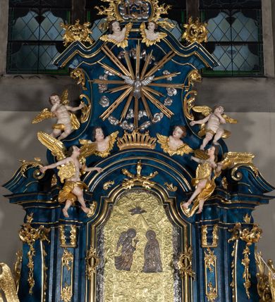 Zdjęcie nr 8: Nastawa architektoniczna, jednoosiowa, jednokondygnacyjna ze zwieńczeniem, ustawiona na wysokim cokole. Ołtarz drewniany, prostopadłościenny, o falistych bokach, mensa drewniana z marmurowym portatylem w drewnianej obudowie. Antepedium ujęte szeroką ramą dekorowaną płaskorzeźbionym motywem wieńca przepasanego wstęgą, w polu medalion z monogramem Marii ujęty wychodzącymi spod jego spodu wiązankami utworzonymi z suchego, wijącego się akantu z karbowaną wstęgą. Nastawa ujęta dwiema parami pilastrów, wewnętrzną z wolutowymi wspornikami w połowie wysokości trzonów oraz zewnętrzną parą o trzonach w formie spływów wolutowych przechodzących w silnie wysunięte do przodu partie cokołu, na których znajdują się postumenty dla figur aniołów. Podpory dźwigają niepełne belkowanie wyłamane na ich osi, nad polem środkowym wyłącznie gzyms przyjmujący kształt łuku półkolistego. Na belkowaniu fragmenty przerwanego, falistego przyczółka oraz para aniołów z kartuszami. W polu środkowym wnęka w formie stojącego prostokąta zamkniętego łukiem segmentowym nadwieszonym z uskokiem, w profilowanej ramie z mniejszym obrazem Matki Boskiej z Dzieciątkiem w kształcie stojącego prostokąta. Poniżej gablota na wota w formie leżącego prostokąta ujęta profilowaną ramą. Kondygnacja flankowana uszami utworzonymi z rocaille&#039;u. Zwieńczenie w formie zbliżonej do kwadratu o wklęsłych bokach przechodzących w woluty, na których siedzą putta, zamknięte fragmentem falistego gzymsu wyłamanego po bokach, na nim rokokowy kartusz z monogramem IHS w polu, flankowany rocaille&#039;em. W polu zwieńczenia rzeźba gołębicy Ducha Świętego w glorii promienistej i otoku z obłoków, w towarzystwie czterech uskrzydlonych główek anielskich. Wszystkie pilastry oraz łuki dekorowane elementami rocaille&#039;u. Struktura w całości polichromowana na sposób imitujący marmoryzację w kolorze niebieskim, ornamentyka i profile złocone, detal srebrzony. 
Wewnątrz skrzyni ołtarzowej napisy „MALOWAŁ LUDWIK WÓJCIK Z KRAKOWA 2 / XI 1914”; „LUDWIK WÓJCIK”; „MALOWALI STOPY”; „23.02.1994 / KONSERWOWALI / KAZIMIERZ PATEJUK / EWA I JERZY SUCHWAŁKO / Z WARSZAWY / PROBOSZCZOWAŁ / KS. BOLESŁAW WAWAK”. 