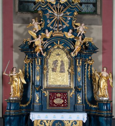 Zdjęcie nr 7: Nastawa architektoniczna, jednoosiowa, jednokondygnacyjna ze zwieńczeniem, ustawiona na wysokim cokole. Ołtarz drewniany, prostopadłościenny, o falistych bokach, mensa drewniana z marmurowym portatylem w drewnianej obudowie. Antepedium ujęte szeroką ramą dekorowaną płaskorzeźbionym motywem wieńca przepasanego wstęgą, w polu medalion z monogramem Marii ujęty wychodzącymi spod jego spodu wiązankami utworzonymi z suchego, wijącego się akantu z karbowaną wstęgą. Nastawa ujęta dwiema parami pilastrów, wewnętrzną z wolutowymi wspornikami w połowie wysokości trzonów oraz zewnętrzną parą o trzonach w formie spływów wolutowych przechodzących w silnie wysunięte do przodu partie cokołu, na których znajdują się postumenty dla figur aniołów. Podpory dźwigają niepełne belkowanie wyłamane na ich osi, nad polem środkowym wyłącznie gzyms przyjmujący kształt łuku półkolistego. Na belkowaniu fragmenty przerwanego, falistego przyczółka oraz para aniołów z kartuszami. W polu środkowym wnęka w formie stojącego prostokąta zamkniętego łukiem segmentowym nadwieszonym z uskokiem, w profilowanej ramie z mniejszym obrazem Matki Boskiej z Dzieciątkiem w kształcie stojącego prostokąta. Poniżej gablota na wota w formie leżącego prostokąta ujęta profilowaną ramą. Kondygnacja flankowana uszami utworzonymi z rocaille&#039;u. Zwieńczenie w formie zbliżonej do kwadratu o wklęsłych bokach przechodzących w woluty, na których siedzą putta, zamknięte fragmentem falistego gzymsu wyłamanego po bokach, na nim rokokowy kartusz z monogramem IHS w polu, flankowany rocaille&#039;em. W polu zwieńczenia rzeźba gołębicy Ducha Świętego w glorii promienistej i otoku z obłoków, w towarzystwie czterech uskrzydlonych główek anielskich. Wszystkie pilastry oraz łuki dekorowane elementami rocaille&#039;u. Struktura w całości polichromowana na sposób imitujący marmoryzację w kolorze niebieskim, ornamentyka i profile złocone, detal srebrzony. 
Wewnątrz skrzyni ołtarzowej napisy „MALOWAŁ LUDWIK WÓJCIK Z KRAKOWA 2 / XI 1914”; „LUDWIK WÓJCIK”; „MALOWALI STOPY”; „23.02.1994 / KONSERWOWALI / KAZIMIERZ PATEJUK / EWA I JERZY SUCHWAŁKO / Z WARSZAWY / PROBOSZCZOWAŁ / KS. BOLESŁAW WAWAK”. 