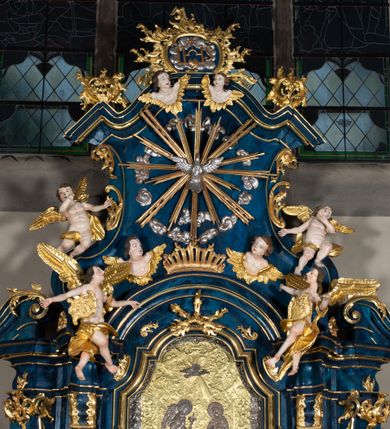 Zdjęcie nr 9: Nastawa architektoniczna, jednoosiowa, jednokondygnacyjna ze zwieńczeniem, ustawiona na wysokim cokole. Ołtarz drewniany, prostopadłościenny, o falistych bokach, mensa drewniana z marmurowym portatylem w drewnianej obudowie. Antepedium ujęte szeroką ramą dekorowaną płaskorzeźbionym motywem wieńca przepasanego wstęgą, w polu medalion z monogramem Marii ujęty wychodzącymi spod jego spodu wiązankami utworzonymi z suchego, wijącego się akantu z karbowaną wstęgą. Nastawa ujęta dwiema parami pilastrów, wewnętrzną z wolutowymi wspornikami w połowie wysokości trzonów oraz zewnętrzną parą o trzonach w formie spływów wolutowych przechodzących w silnie wysunięte do przodu partie cokołu, na których znajdują się postumenty dla figur aniołów. Podpory dźwigają niepełne belkowanie wyłamane na ich osi, nad polem środkowym wyłącznie gzyms przyjmujący kształt łuku półkolistego. Na belkowaniu fragmenty przerwanego, falistego przyczółka oraz para aniołów z kartuszami. W polu środkowym wnęka w formie stojącego prostokąta zamkniętego łukiem segmentowym nadwieszonym z uskokiem, w profilowanej ramie z mniejszym obrazem Matki Boskiej z Dzieciątkiem w kształcie stojącego prostokąta. Poniżej gablota na wota w formie leżącego prostokąta ujęta profilowaną ramą. Kondygnacja flankowana uszami utworzonymi z rocaille'u. Zwieńczenie w formie zbliżonej do kwadratu o wklęsłych bokach przechodzących w woluty, na których siedzą putta, zamknięte fragmentem falistego gzymsu wyłamanego po bokach, na nim rokokowy kartusz z monogramem IHS w polu, flankowany rocaille'em. W polu zwieńczenia rzeźba gołębicy Ducha Świętego w glorii promienistej i otoku z obłoków, w towarzystwie czterech uskrzydlonych główek anielskich. Wszystkie pilastry oraz łuki dekorowane elementami rocaille'u. Struktura w całości polichromowana na sposób imitujący marmoryzację w kolorze niebieskim, ornamentyka i profile złocone, detal srebrzony. 
Wewnątrz skrzyni ołtarzowej napisy „MALOWAŁ LUDWIK WÓJCIK Z KRAKOWA 2 / XI 1914”; „LUDWIK WÓJCIK”; „MALOWALI STOPY”; „23.02.1994 / KONSERWOWALI / KAZIMIERZ PATEJUK / EWA I JERZY SUCHWAŁKO / Z WARSZAWY / PROBOSZCZOWAŁ / KS. BOLESŁAW WAWAK”. 