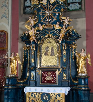 Zdjęcie nr 6: Nastawa architektoniczna, jednoosiowa, jednokondygnacyjna ze zwieńczeniem, ustawiona na wysokim cokole. Ołtarz drewniany, prostopadłościenny, o falistych bokach, mensa drewniana z marmurowym portatylem w drewnianej obudowie. Antepedium ujęte szeroką ramą dekorowaną płaskorzeźbionym motywem wieńca przepasanego wstęgą, w polu medalion z monogramem Marii ujęty wychodzącymi spod jego spodu wiązankami utworzonymi z suchego, wijącego się akantu z karbowaną wstęgą. Nastawa ujęta dwiema parami pilastrów, wewnętrzną z wolutowymi wspornikami w połowie wysokości trzonów oraz zewnętrzną parą o trzonach w formie spływów wolutowych przechodzących w silnie wysunięte do przodu partie cokołu, na których znajdują się postumenty dla figur aniołów. Podpory dźwigają niepełne belkowanie wyłamane na ich osi, nad polem środkowym wyłącznie gzyms przyjmujący kształt łuku półkolistego. Na belkowaniu fragmenty przerwanego, falistego przyczółka oraz para aniołów z kartuszami. W polu środkowym wnęka w formie stojącego prostokąta zamkniętego łukiem segmentowym nadwieszonym z uskokiem, w profilowanej ramie z mniejszym obrazem Matki Boskiej z Dzieciątkiem w kształcie stojącego prostokąta. Poniżej gablota na wota w formie leżącego prostokąta ujęta profilowaną ramą. Kondygnacja flankowana uszami utworzonymi z rocaille'u. Zwieńczenie w formie zbliżonej do kwadratu o wklęsłych bokach przechodzących w woluty, na których siedzą putta, zamknięte fragmentem falistego gzymsu wyłamanego po bokach, na nim rokokowy kartusz z monogramem IHS w polu, flankowany rocaille'em. W polu zwieńczenia rzeźba gołębicy Ducha Świętego w glorii promienistej i otoku z obłoków, w towarzystwie czterech uskrzydlonych główek anielskich. Wszystkie pilastry oraz łuki dekorowane elementami rocaille'u. Struktura w całości polichromowana na sposób imitujący marmoryzację w kolorze niebieskim, ornamentyka i profile złocone, detal srebrzony. 
Wewnątrz skrzyni ołtarzowej napisy „MALOWAŁ LUDWIK WÓJCIK Z KRAKOWA 2 / XI 1914”; „LUDWIK WÓJCIK”; „MALOWALI STOPY”; „23.02.1994 / KONSERWOWALI / KAZIMIERZ PATEJUK / EWA I JERZY SUCHWAŁKO / Z WARSZAWY / PROBOSZCZOWAŁ / KS. BOLESŁAW WAWAK”. 