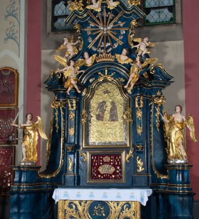 Zdjęcie nr 5: Nastawa architektoniczna, jednoosiowa, jednokondygnacyjna ze zwieńczeniem, ustawiona na wysokim cokole. Ołtarz drewniany, prostopadłościenny, o falistych bokach, mensa drewniana z marmurowym portatylem w drewnianej obudowie. Antepedium ujęte szeroką ramą dekorowaną płaskorzeźbionym motywem wieńca przepasanego wstęgą, w polu medalion z monogramem Marii ujęty wychodzącymi spod jego spodu wiązankami utworzonymi z suchego, wijącego się akantu z karbowaną wstęgą. Nastawa ujęta dwiema parami pilastrów, wewnętrzną z wolutowymi wspornikami w połowie wysokości trzonów oraz zewnętrzną parą o trzonach w formie spływów wolutowych przechodzących w silnie wysunięte do przodu partie cokołu, na których znajdują się postumenty dla figur aniołów. Podpory dźwigają niepełne belkowanie wyłamane na ich osi, nad polem środkowym wyłącznie gzyms przyjmujący kształt łuku półkolistego. Na belkowaniu fragmenty przerwanego, falistego przyczółka oraz para aniołów z kartuszami. W polu środkowym wnęka w formie stojącego prostokąta zamkniętego łukiem segmentowym nadwieszonym z uskokiem, w profilowanej ramie z mniejszym obrazem Matki Boskiej z Dzieciątkiem w kształcie stojącego prostokąta. Poniżej gablota na wota w formie leżącego prostokąta ujęta profilowaną ramą. Kondygnacja flankowana uszami utworzonymi z rocaille'u. Zwieńczenie w formie zbliżonej do kwadratu o wklęsłych bokach przechodzących w woluty, na których siedzą putta, zamknięte fragmentem falistego gzymsu wyłamanego po bokach, na nim rokokowy kartusz z monogramem IHS w polu, flankowany rocaille'em. W polu zwieńczenia rzeźba gołębicy Ducha Świętego w glorii promienistej i otoku z obłoków, w towarzystwie czterech uskrzydlonych główek anielskich. Wszystkie pilastry oraz łuki dekorowane elementami rocaille'u. Struktura w całości polichromowana na sposób imitujący marmoryzację w kolorze niebieskim, ornamentyka i profile złocone, detal srebrzony. 
Wewnątrz skrzyni ołtarzowej napisy „MALOWAŁ LUDWIK WÓJCIK Z KRAKOWA 2 / XI 1914”; „LUDWIK WÓJCIK”; „MALOWALI STOPY”; „23.02.1994 / KONSERWOWALI / KAZIMIERZ PATEJUK / EWA I JERZY SUCHWAŁKO / Z WARSZAWY / PROBOSZCZOWAŁ / KS. BOLESŁAW WAWAK”. 