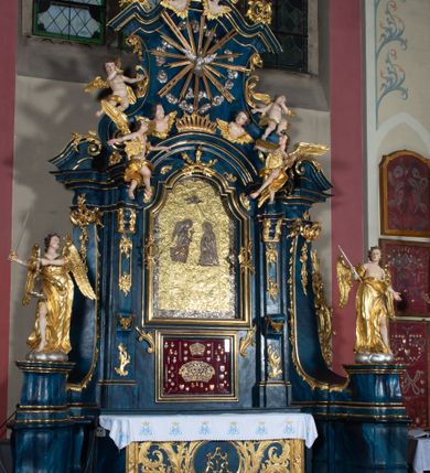 Zdjęcie nr 4: Nastawa architektoniczna, jednoosiowa, jednokondygnacyjna ze zwieńczeniem, ustawiona na wysokim cokole. Ołtarz drewniany, prostopadłościenny, o falistych bokach, mensa drewniana z marmurowym portatylem w drewnianej obudowie. Antepedium ujęte szeroką ramą dekorowaną płaskorzeźbionym motywem wieńca przepasanego wstęgą, w polu medalion z monogramem Marii ujęty wychodzącymi spod jego spodu wiązankami utworzonymi z suchego, wijącego się akantu z karbowaną wstęgą. Nastawa ujęta dwiema parami pilastrów, wewnętrzną z wolutowymi wspornikami w połowie wysokości trzonów oraz zewnętrzną parą o trzonach w formie spływów wolutowych przechodzących w silnie wysunięte do przodu partie cokołu, na których znajdują się postumenty dla figur aniołów. Podpory dźwigają niepełne belkowanie wyłamane na ich osi, nad polem środkowym wyłącznie gzyms przyjmujący kształt łuku półkolistego. Na belkowaniu fragmenty przerwanego, falistego przyczółka oraz para aniołów z kartuszami. W polu środkowym wnęka w formie stojącego prostokąta zamkniętego łukiem segmentowym nadwieszonym z uskokiem, w profilowanej ramie z mniejszym obrazem Matki Boskiej z Dzieciątkiem w kształcie stojącego prostokąta. Poniżej gablota na wota w formie leżącego prostokąta ujęta profilowaną ramą. Kondygnacja flankowana uszami utworzonymi z rocaille'u. Zwieńczenie w formie zbliżonej do kwadratu o wklęsłych bokach przechodzących w woluty, na których siedzą putta, zamknięte fragmentem falistego gzymsu wyłamanego po bokach, na nim rokokowy kartusz z monogramem IHS w polu, flankowany rocaille'em. W polu zwieńczenia rzeźba gołębicy Ducha Świętego w glorii promienistej i otoku z obłoków, w towarzystwie czterech uskrzydlonych główek anielskich. Wszystkie pilastry oraz łuki dekorowane elementami rocaille'u. Struktura w całości polichromowana na sposób imitujący marmoryzację w kolorze niebieskim, ornamentyka i profile złocone, detal srebrzony. 
Wewnątrz skrzyni ołtarzowej napisy „MALOWAŁ LUDWIK WÓJCIK Z KRAKOWA 2 / XI 1914”; „LUDWIK WÓJCIK”; „MALOWALI STOPY”; „23.02.1994 / KONSERWOWALI / KAZIMIERZ PATEJUK / EWA I JERZY SUCHWAŁKO / Z WARSZAWY / PROBOSZCZOWAŁ / KS. BOLESŁAW WAWAK”. 
