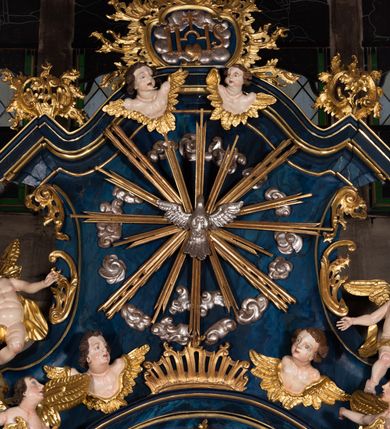 Zdjęcie nr 13: Nastawa architektoniczna, jednoosiowa, jednokondygnacyjna ze zwieńczeniem, ustawiona na wysokim cokole. Ołtarz drewniany, prostopadłościenny, o falistych bokach, mensa drewniana z marmurowym portatylem w drewnianej obudowie. Antepedium ujęte szeroką ramą dekorowaną płaskorzeźbionym motywem wieńca przepasanego wstęgą, w polu medalion z monogramem Marii ujęty wychodzącymi spod jego spodu wiązankami utworzonymi z suchego, wijącego się akantu z karbowaną wstęgą. Nastawa ujęta dwiema parami pilastrów, wewnętrzną z wolutowymi wspornikami w połowie wysokości trzonów oraz zewnętrzną parą o trzonach w formie spływów wolutowych przechodzących w silnie wysunięte do przodu partie cokołu, na których znajdują się postumenty dla figur aniołów. Podpory dźwigają niepełne belkowanie wyłamane na ich osi, nad polem środkowym wyłącznie gzyms przyjmujący kształt łuku półkolistego. Na belkowaniu fragmenty przerwanego, falistego przyczółka oraz para aniołów z kartuszami. W polu środkowym wnęka w formie stojącego prostokąta zamkniętego łukiem segmentowym nadwieszonym z uskokiem, w profilowanej ramie z mniejszym obrazem Matki Boskiej z Dzieciątkiem w kształcie stojącego prostokąta. Poniżej gablota na wota w formie leżącego prostokąta ujęta profilowaną ramą. Kondygnacja flankowana uszami utworzonymi z rocaille'u. Zwieńczenie w formie zbliżonej do kwadratu o wklęsłych bokach przechodzących w woluty, na których siedzą putta, zamknięte fragmentem falistego gzymsu wyłamanego po bokach, na nim rokokowy kartusz z monogramem IHS w polu, flankowany rocaille'em. W polu zwieńczenia rzeźba gołębicy Ducha Świętego w glorii promienistej i otoku z obłoków, w towarzystwie czterech uskrzydlonych główek anielskich. Wszystkie pilastry oraz łuki dekorowane elementami rocaille'u. Struktura w całości polichromowana na sposób imitujący marmoryzację w kolorze niebieskim, ornamentyka i profile złocone, detal srebrzony. 
Wewnątrz skrzyni ołtarzowej napisy „MALOWAŁ LUDWIK WÓJCIK Z KRAKOWA 2 / XI 1914”; „LUDWIK WÓJCIK”; „MALOWALI STOPY”; „23.02.1994 / KONSERWOWALI / KAZIMIERZ PATEJUK / EWA I JERZY SUCHWAŁKO / Z WARSZAWY / PROBOSZCZOWAŁ / KS. BOLESŁAW WAWAK”. 