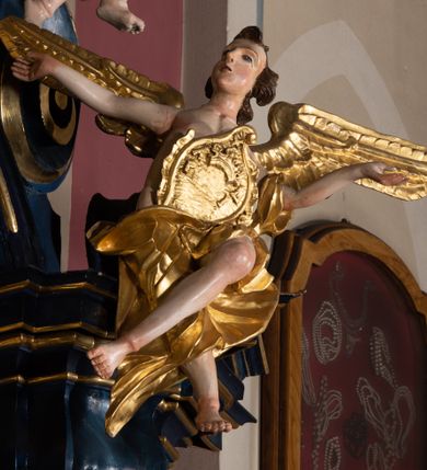 Zdjęcie nr 18: Nastawa architektoniczna, jednoosiowa, jednokondygnacyjna ze zwieńczeniem, ustawiona na wysokim cokole. Ołtarz drewniany, prostopadłościenny, o falistych bokach, mensa drewniana z marmurowym portatylem w drewnianej obudowie. Antepedium ujęte szeroką ramą dekorowaną płaskorzeźbionym motywem wieńca przepasanego wstęgą, w polu medalion z monogramem Marii ujęty wychodzącymi spod jego spodu wiązankami utworzonymi z suchego, wijącego się akantu z karbowaną wstęgą. Nastawa ujęta dwiema parami pilastrów, wewnętrzną z wolutowymi wspornikami w połowie wysokości trzonów oraz zewnętrzną parą o trzonach w formie spływów wolutowych przechodzących w silnie wysunięte do przodu partie cokołu, na których znajdują się postumenty dla figur aniołów. Podpory dźwigają niepełne belkowanie wyłamane na ich osi, nad polem środkowym wyłącznie gzyms przyjmujący kształt łuku półkolistego. Na belkowaniu fragmenty przerwanego, falistego przyczółka oraz para aniołów z kartuszami. W polu środkowym wnęka w formie stojącego prostokąta zamkniętego łukiem segmentowym nadwieszonym z uskokiem, w profilowanej ramie z mniejszym obrazem Matki Boskiej z Dzieciątkiem w kształcie stojącego prostokąta. Poniżej gablota na wota w formie leżącego prostokąta ujęta profilowaną ramą. Kondygnacja flankowana uszami utworzonymi z rocaille&#039;u. Zwieńczenie w formie zbliżonej do kwadratu o wklęsłych bokach przechodzących w woluty, na których siedzą putta, zamknięte fragmentem falistego gzymsu wyłamanego po bokach, na nim rokokowy kartusz z monogramem IHS w polu, flankowany rocaille&#039;em. W polu zwieńczenia rzeźba gołębicy Ducha Świętego w glorii promienistej i otoku z obłoków, w towarzystwie czterech uskrzydlonych główek anielskich. Wszystkie pilastry oraz łuki dekorowane elementami rocaille&#039;u. Struktura w całości polichromowana na sposób imitujący marmoryzację w kolorze niebieskim, ornamentyka i profile złocone, detal srebrzony. 
Wewnątrz skrzyni ołtarzowej napisy „MALOWAŁ LUDWIK WÓJCIK Z KRAKOWA 2 / XI 1914”; „LUDWIK WÓJCIK”; „MALOWALI STOPY”; „23.02.1994 / KONSERWOWALI / KAZIMIERZ PATEJUK / EWA I JERZY SUCHWAŁKO / Z WARSZAWY / PROBOSZCZOWAŁ / KS. BOLESŁAW WAWAK”. 