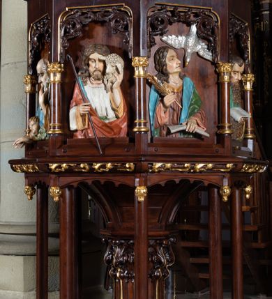 Zdjęcie nr 2: Ambona wolnostojąca, ustawiona na ośmiobocznej, dwustopniowej podstawie oraz środkowej kolumnie dekorowanej liśćmi akantu i otaczającymi ją ośmioma kolumienkami. Kosz na planie ośmioboku, na niewielkiej podstawie z podwieszonymi kwiatonami, z dostępem w postaci jednobiegowych schodów z dwiema balustradami dekorowanymi stylizowanymi blendami. Ścianki kosza wydzielone niskimi kolumienkami. W polach prostokątne nisze w formie stojącego prostokąta, w górnej partii dekorowane stylizowanym astwerkiem, z płaskorzeźbionymi czterech ewangelistów i Chrystusa Dobrego Pasterza. Parapet w formie gzymsu z pulpitem. Struktura politurowana w kolorze ciemnego brązu, detal złocony, wizerunki postaci polichromowane. Płaskorzeźby Chrystusa i ewangelistów ukazane w półpostaci. Apostołowie zwróceni są w trzech czwartych, w dłoniach trzymają pióra i zwoje lub księgi, towarzyszą im ich symbole. Chrystus lewą ręką podtrzymuje na ramieniu owcę, w prawej trzyma laskę pasterską. Wszyscy ubrani są w obszerne tuniki i płaszcze narzucone na ramiona. 