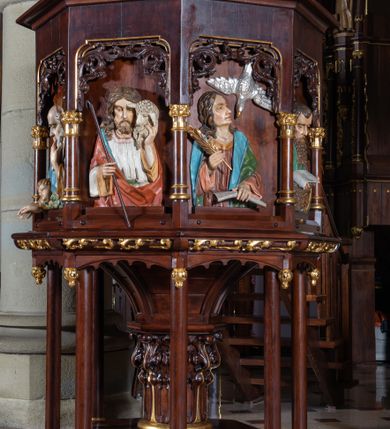 Zdjęcie nr 3: Ambona wolnostojąca, ustawiona na ośmiobocznej, dwustopniowej podstawie oraz środkowej kolumnie dekorowanej liśćmi akantu i otaczającymi ją ośmioma kolumienkami. Kosz na planie ośmioboku, na niewielkiej podstawie z podwieszonymi kwiatonami, z dostępem w postaci jednobiegowych schodów z dwiema balustradami dekorowanymi stylizowanymi blendami. Ścianki kosza wydzielone niskimi kolumienkami. W polach prostokątne nisze w formie stojącego prostokąta, w górnej partii dekorowane stylizowanym astwerkiem, z płaskorzeźbionymi czterech ewangelistów i Chrystusa Dobrego Pasterza. Parapet w formie gzymsu z pulpitem. Struktura politurowana w kolorze ciemnego brązu, detal złocony, wizerunki postaci polichromowane. Płaskorzeźby Chrystusa i ewangelistów ukazane w półpostaci. Apostołowie zwróceni są w trzech czwartych, w dłoniach trzymają pióra i zwoje lub księgi, towarzyszą im ich symbole. Chrystus lewą ręką podtrzymuje na ramieniu owcę, w prawej trzyma laskę pasterską. Wszyscy ubrani są w obszerne tuniki i płaszcze narzucone na ramiona. 