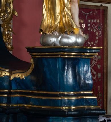 Zdjęcie nr 5: Rzeźba ścięta z tyłu, drążona, ustawiona na podstawie w formie obłoku. Figura zwrócona frontalnie, z głową skierowaną w prawo, w kontrapoście, z rozłożonymi na boki rękami, w tym z prawą uniesioną, z berłem w dłoni. Twarz o młodzieńczych rysach, pociągła, o oczach osadzonych blisko siebie, prostym nosie i lekko rozchylonych ustach, okolona krótkimi włosami rzeźbionymi w pojedyncze, grube i rozwiane pukle. Anioł ubrany w długą suknię, odsłaniającą połowę torsu oraz lewą nogę, przepasaną w talii. Tkanina zawinięta wokół ramion i torsu obficie drapowana, przylegająca do ciała w partii bioder, dołem rozwiana. Skrzydła duże, złożone po bokach postaci. Polichromia naturalistyczna w odsłoniętych partiach ciała, szaty i skrzydła złocone, berło i obłok srebrzone.