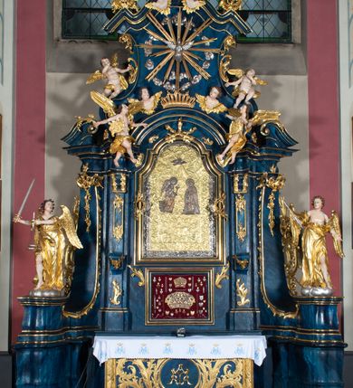 Zdjęcie nr 6: Rzeźba ścięta z tyłu, drążona, ustawiona na podstawie w formie obłoku. Figura zwrócona frontalnie, z głową skierowaną w prawo, w kontrapoście, z rozłożonymi na boki rękami, w tym z prawą uniesioną, z berłem w dłoni. Twarz o młodzieńczych rysach, pociągła, o oczach osadzonych blisko siebie, prostym nosie i lekko rozchylonych ustach, okolona krótkimi włosami rzeźbionymi w pojedyncze, grube i rozwiane pukle. Anioł ubrany w długą suknię, odsłaniającą połowę torsu oraz lewą nogę, przepasaną w talii. Tkanina zawinięta wokół ramion i torsu obficie drapowana, przylegająca do ciała w partii bioder, dołem rozwiana. Skrzydła duże, złożone po bokach postaci. Polichromia naturalistyczna w odsłoniętych partiach ciała, szaty i skrzydła złocone, berło i obłok srebrzone.