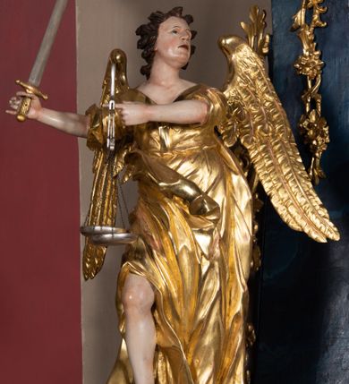 Zdjęcie nr 2: Rzeźba ścięta z tyłu, drążona, ustawiona na podstawie w formie obłoku. Figura zwrócona frontalnie, w kontrapoście, z uniesioną głową skierowaną w lewo oraz uniesionymi rękami wyciągniętymi w przeciwną stroną. Na wyciągniętym palcu wskazującym lewej dłoni zawieszona waga szalkowa, prawa dzierży miecz skierowany ostrzem w górę. Twarz pociągła, o młodzieńczych rysach, migdałowate oczy usytuowane blisko siebie, skierowane do góry, nos prosty, usta rozchylone; włosy krótkie, rzeźbione w grube, pojedyncze i rozwiane pukle. Postać ubrana w długą tunikę z szerokimi, krótkimi rękawami, przepasaną w talii, odsłaniającą prawą nogę oraz w płaszcz o wywiniętym brzegu, przerzucony z lewego boku postaci. Skrzydła duże, złożone po bokach postaci. Polichromia naturalistyczna w odsłoniętych partiach ciała, szaty złocone, atrybuty i obłok srebrzone.