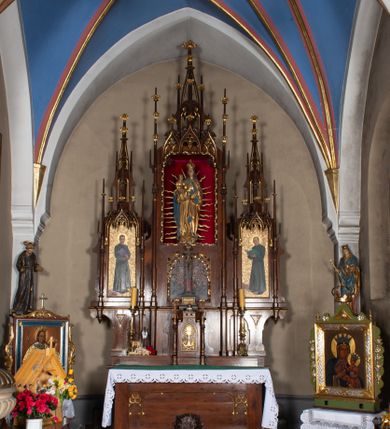 Zdjęcie nr 5: Nastawa ołtarzowa architektoniczna, trójosiowa, jednokondygnacyjna, z predellą. Ołtarz drewniany prostopadłościenny, z drewnianą mensą i portatylem z marmuru dębnickiego. W polu antepedium płycina ujęta blendami i maswerkiem; w centrum monogram Marii w wieńcu róż. Tabernakulum w formie prostopadłościennej skrzynki zamkniętej krenelażem, ujęte dwiema kolumienkami z pinaklami, z prostokątnymi drzwiczkami zamkniętymi łukiem ostrym z płaskorzeźbionym przedstawieniem kielicha, hostią w glorii, winną latoroślą i kłosami zbóż. Osie nastawy wydzielone smukłymi półkolumnami, zakończonymi pinaklami z kwiatonami. Oś środkowa dwukrotnie wyższa i szersza od bocznych. W polu środkowym nisza w formie stojącego prostokąta z figurą Matki Boskiej z Dzieciątkiem. Wnęka ujęta ramą dekorowaną ornamentem roślinnym. Poniżej płycina w kształcie kwadratu zamkniętego łukiem segmentowym, ujęta maswerkową ramą z przedstawieniem Chrystusa z więźniami obozu koncentracyjnego i duchownymi. Oś główna zamknięta baldachimem utworzonym z trzech wimperg oraz szczytem z pinakli i kwiatonów. Osie boczne z wnękami z obrazami bł. Stanisława Pyrtka i bł. Piotra Dańkowskiego, zwieńczonymi baldachimami utworzonymi z trzech wimperg i zakończonymi gotyckimi wieżyczkami. Struktura nastawy politurowana w kolorze jasnobrązowym, ornamentyka i detale architektoniczne złocone. 

