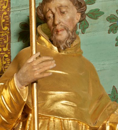 Zdjęcie nr 2: Rzeźba ścięta z tyłu, drążona, ustawiona na niskiej podstawie. Święty ukazany w całej postaci, frontalnie, w kontrapoście, o lekko przechylonej sylwetce, z delikatnie pochyloną głową, skierowaną w prawo; w prawej, ugiętej ręce trzyma laskę zakończoną krzyżem, lewa ma uniesioną i ugiętą. Twarz ma szczupłą, pociągłą, o wyraźnie rzeźbionych, ściągniętych łukach brwiowych, głęboko osadzonych oczach skierowanych w dół, prostym nosie i pełnych ustach; okoloną krótką kręconą brodą oraz krótkimi włosami, układającymi się w grube pukle, na głowie tonsura. Ubrany jest w złocony habit, przewiązany w pasie sznurem z trzema guzami, przylegający ścisłe do ciała, obficie fałdowany w grube, głęboko rzeźbione fałdy. Na ramiona ma narzuconą złoconą pelerynkę z kapturem, dłuższą na plecach, delikatnie marszczoną na piersi. Polichromia ciała figury naturalistyczna. 