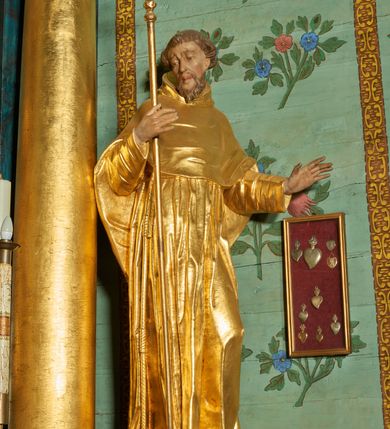 Zdjęcie nr 4: Rzeźba ścięta z tyłu, drążona, ustawiona na niskiej podstawie. Święty ukazany w całej postaci, frontalnie, w kontrapoście, o lekko przechylonej sylwetce, z delikatnie pochyloną głową, skierowaną w prawo; w prawej, ugiętej ręce trzyma laskę zakończoną krzyżem, lewa ma uniesioną i ugiętą. Twarz ma szczupłą, pociągłą, o wyraźnie rzeźbionych, ściągniętych łukach brwiowych, głęboko osadzonych oczach skierowanych w dół, prostym nosie i pełnych ustach; okoloną krótką kręconą brodą oraz krótkimi włosami, układającymi się w grube pukle, na głowie tonsura. Ubrany jest w złocony habit, przewiązany w pasie sznurem z trzema guzami, przylegający ścisłe do ciała, obficie fałdowany w grube, głęboko rzeźbione fałdy. Na ramiona ma narzuconą złoconą pelerynkę z kapturem, dłuższą na plecach, delikatnie marszczoną na piersi. Polichromia ciała figury naturalistyczna. 