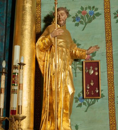 Zdjęcie nr 5: Rzeźba ścięta z tyłu, drążona, ustawiona na niskiej podstawie. Święty ukazany w całej postaci, frontalnie, w kontrapoście, o lekko przechylonej sylwetce, z delikatnie pochyloną głową, skierowaną w prawo; w prawej, ugiętej ręce trzyma laskę zakończoną krzyżem, lewa ma uniesioną i ugiętą. Twarz ma szczupłą, pociągłą, o wyraźnie rzeźbionych, ściągniętych łukach brwiowych, głęboko osadzonych oczach skierowanych w dół, prostym nosie i pełnych ustach; okoloną krótką kręconą brodą oraz krótkimi włosami, układającymi się w grube pukle, na głowie tonsura. Ubrany jest w złocony habit, przewiązany w pasie sznurem z trzema guzami, przylegający ścisłe do ciała, obficie fałdowany w grube, głęboko rzeźbione fałdy. Na ramiona ma narzuconą złoconą pelerynkę z kapturem, dłuższą na plecach, delikatnie marszczoną na piersi. Polichromia ciała figury naturalistyczna. 
