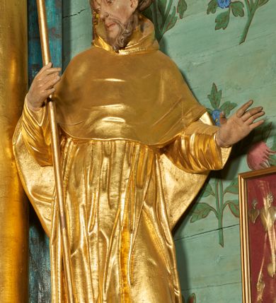 Zdjęcie nr 3: Rzeźba ścięta z tyłu, drążona, ustawiona na niskiej podstawie. Święty ukazany w całej postaci, frontalnie, w kontrapoście, o lekko przechylonej sylwetce, z delikatnie pochyloną głową, skierowaną w prawo; w prawej, ugiętej ręce trzyma laskę zakończoną krzyżem, lewa ma uniesioną i ugiętą. Twarz ma szczupłą, pociągłą, o wyraźnie rzeźbionych, ściągniętych łukach brwiowych, głęboko osadzonych oczach skierowanych w dół, prostym nosie i pełnych ustach; okoloną krótką kręconą brodą oraz krótkimi włosami, układającymi się w grube pukle, na głowie tonsura. Ubrany jest w złocony habit, przewiązany w pasie sznurem z trzema guzami, przylegający ścisłe do ciała, obficie fałdowany w grube, głęboko rzeźbione fałdy. Na ramiona ma narzuconą złoconą pelerynkę z kapturem, dłuższą na plecach, delikatnie marszczoną na piersi. Polichromia ciała figury naturalistyczna. 