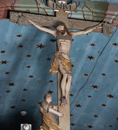 Zdjęcie nr 9: Belka tęczowa o falistym zarysie, zwieńczona profilowanym gzymsem. Z przodu napis „WSZYSCY KTÓRZY PRZYCHO/DZICIE OBACZCIE I UWAŻCIE: / OCZY MOJE BĘDĄ OTWARTE I USZY NACHYLONE NA PROŚBĘ TEGO KTÓRY SIĘ NA TEM MIEJSCU MODLIĆ BĘDZIE.”, na odwrociu napis „TEN KOŚCIÓŁ FUNDOWANY/ PRZEZ BARBARĘ LISICKĄ. / KONSEKROWANY W ROKU 1766. / ODNOMIONY W ROKU 1902. PO RAZ DRUGI W 1980”. Na belce ustawione figury ze sceny Ukrzyżowania. Pośrodku krzyż, do którego przybita jest trzema gwoździami figura umarłego Chrystusa. Ciało w lekkim zwisie o horyzontalnie rozłożonych ramionach, z głową opadającą na prawe ramię, z lekko ugiętymi nogami oraz stopami skrzyżowanymi w układzie prawa na lewą. Twarz ma szczupłą, o zamkniętych oczach, uniesionych brwiach, dużym, wąskim nosie oraz otwartych ustach, okoloną krótką, falowaną brodą i silnie kręconymi włosami opadającymi na ramiona. Na głowie ma koronę cierniową. Ciało jest wychudzone, o silnie zaznaczonych, guzowatych żebrach klatki piersiowej oraz mięśniach i ścięgnach ramion i ud. W partii bioder złocone perizonium zawieszone na sznurze, odsłaniające prawe udo, zawiązane ze zwisem tkaniny na prawym biodrze. 
U dołu krzyża klęczy Maria Magdalena, zwrócona w trzech czwartych w lewo, z uniesioną głową, obiema rękami obejmuje belkę. Twarz ma pociągłą, o małych oczach, krótkim nosie i wąskich ustach, okoloną długimi falowanymi włosami opadającymi na ramiona. Ubrana jest w srebrzoną suknię oraz złocony płaszcz. 
Po prawej stronie krzyża figura Marii, zwrócona w trzech czwartych w lewo, w kontrapoście, z pochyloną głową i rękami skrzyżowanymi na piersi. Twarz ma pociągłą, o wąskich oczach, uniesionych brwiach, małych nosie i ustach. Ubrana jest w srebrzoną suknię oraz złocony płaszcz narzucony na głowę i ramiona, wokół głowy ma białą podwikę. 
Po lewej stronie krzyża św. Jan zwrócony w trzech czwartych w prawo, w kontrapoście, prawą rękę kładzie na piersi, w lewej, opuszczonej trzyma księgę. Twarz ma pociągłą, o wąskich oczach, małych nosie i ustach, okoloną brązowymi, silnie kręconymi włosami, sięgającymi ramion, z czubem nad czołem. Ubrany jest w srebrzoną tunikę z kołnierzykiem, przepasaną w talii oraz złocony płaszcz zarzucony na prawe ramię i podtrzymywany na lewym boku. Polichromia ciał postaci naturalistyczna. 