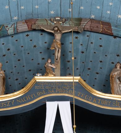 Zdjęcie nr 2: Belka tęczowa o falistym zarysie, zwieńczona profilowanym gzymsem. Z przodu napis „WSZYSCY KTÓRZY PRZYCHO/DZICIE OBACZCIE I UWAŻCIE: / OCZY MOJE BĘDĄ OTWARTE I USZY NACHYLONE NA PROŚBĘ TEGO KTÓRY SIĘ NA TEM MIEJSCU MODLIĆ BĘDZIE.”, na odwrociu napis „TEN KOŚCIÓŁ FUNDOWANY/ PRZEZ BARBARĘ LISICKĄ. / KONSEKROWANY W ROKU 1766. / ODNOMIONY W ROKU 1902. PO RAZ DRUGI W 1980”. Na belce ustawione figury ze sceny Ukrzyżowania. Pośrodku krzyż, do którego przybita jest trzema gwoździami figura umarłego Chrystusa. Ciało w lekkim zwisie o horyzontalnie rozłożonych ramionach, z głową opadającą na prawe ramię, z lekko ugiętymi nogami oraz stopami skrzyżowanymi w układzie prawa na lewą. Twarz ma szczupłą, o zamkniętych oczach, uniesionych brwiach, dużym, wąskim nosie oraz otwartych ustach, okoloną krótką, falowaną brodą i silnie kręconymi włosami opadającymi na ramiona. Na głowie ma koronę cierniową. Ciało jest wychudzone, o silnie zaznaczonych, guzowatych żebrach klatki piersiowej oraz mięśniach i ścięgnach ramion i ud. W partii bioder złocone perizonium zawieszone na sznurze, odsłaniające prawe udo, zawiązane ze zwisem tkaniny na prawym biodrze. 
U dołu krzyża klęczy Maria Magdalena, zwrócona w trzech czwartych w lewo, z uniesioną głową, obiema rękami obejmuje belkę. Twarz ma pociągłą, o małych oczach, krótkim nosie i wąskich ustach, okoloną długimi falowanymi włosami opadającymi na ramiona. Ubrana jest w srebrzoną suknię oraz złocony płaszcz. 
Po prawej stronie krzyża figura Marii, zwrócona w trzech czwartych w lewo, w kontrapoście, z pochyloną głową i rękami skrzyżowanymi na piersi. Twarz ma pociągłą, o wąskich oczach, uniesionych brwiach, małych nosie i ustach. Ubrana jest w srebrzoną suknię oraz złocony płaszcz narzucony na głowę i ramiona, wokół głowy ma białą podwikę. 
Po lewej stronie krzyża św. Jan zwrócony w trzech czwartych w prawo, w kontrapoście, prawą rękę kładzie na piersi, w lewej, opuszczonej trzyma księgę. Twarz ma pociągłą, o wąskich oczach, małych nosie i ustach, okoloną brązowymi, silnie kręconymi włosami, sięgającymi ramion, z czubem nad czołem. Ubrany jest w srebrzoną tunikę z kołnierzykiem, przepasaną w talii oraz złocony płaszcz zarzucony na prawe ramię i podtrzymywany na lewym boku. Polichromia ciał postaci naturalistyczna. 