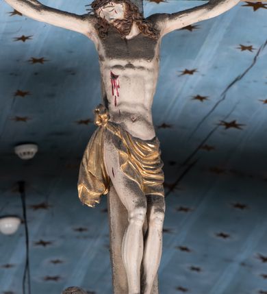 Zdjęcie nr 11: Belka tęczowa o falistym zarysie, zwieńczona profilowanym gzymsem. Z przodu napis „WSZYSCY KTÓRZY PRZYCHO/DZICIE OBACZCIE I UWAŻCIE: / OCZY MOJE BĘDĄ OTWARTE I USZY NACHYLONE NA PROŚBĘ TEGO KTÓRY SIĘ NA TEM MIEJSCU MODLIĆ BĘDZIE.”, na odwrociu napis „TEN KOŚCIÓŁ FUNDOWANY/ PRZEZ BARBARĘ LISICKĄ. / KONSEKROWANY W ROKU 1766. / ODNOMIONY W ROKU 1902. PO RAZ DRUGI W 1980”. Na belce ustawione figury ze sceny Ukrzyżowania. Pośrodku krzyż, do którego przybita jest trzema gwoździami figura umarłego Chrystusa. Ciało w lekkim zwisie o horyzontalnie rozłożonych ramionach, z głową opadającą na prawe ramię, z lekko ugiętymi nogami oraz stopami skrzyżowanymi w układzie prawa na lewą. Twarz ma szczupłą, o zamkniętych oczach, uniesionych brwiach, dużym, wąskim nosie oraz otwartych ustach, okoloną krótką, falowaną brodą i silnie kręconymi włosami opadającymi na ramiona. Na głowie ma koronę cierniową. Ciało jest wychudzone, o silnie zaznaczonych, guzowatych żebrach klatki piersiowej oraz mięśniach i ścięgnach ramion i ud. W partii bioder złocone perizonium zawieszone na sznurze, odsłaniające prawe udo, zawiązane ze zwisem tkaniny na prawym biodrze. 
U dołu krzyża klęczy Maria Magdalena, zwrócona w trzech czwartych w lewo, z uniesioną głową, obiema rękami obejmuje belkę. Twarz ma pociągłą, o małych oczach, krótkim nosie i wąskich ustach, okoloną długimi falowanymi włosami opadającymi na ramiona. Ubrana jest w srebrzoną suknię oraz złocony płaszcz. 
Po prawej stronie krzyża figura Marii, zwrócona w trzech czwartych w lewo, w kontrapoście, z pochyloną głową i rękami skrzyżowanymi na piersi. Twarz ma pociągłą, o wąskich oczach, uniesionych brwiach, małych nosie i ustach. Ubrana jest w srebrzoną suknię oraz złocony płaszcz narzucony na głowę i ramiona, wokół głowy ma białą podwikę. 
Po lewej stronie krzyża św. Jan zwrócony w trzech czwartych w prawo, w kontrapoście, prawą rękę kładzie na piersi, w lewej, opuszczonej trzyma księgę. Twarz ma pociągłą, o wąskich oczach, małych nosie i ustach, okoloną brązowymi, silnie kręconymi włosami, sięgającymi ramion, z czubem nad czołem. Ubrany jest w srebrzoną tunikę z kołnierzykiem, przepasaną w talii oraz złocony płaszcz zarzucony na prawe ramię i podtrzymywany na lewym boku. Polichromia ciał postaci naturalistyczna. 
