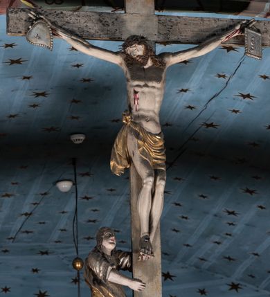 Zdjęcie nr 10: Belka tęczowa o falistym zarysie, zwieńczona profilowanym gzymsem. Z przodu napis „WSZYSCY KTÓRZY PRZYCHO/DZICIE OBACZCIE I UWAŻCIE: / OCZY MOJE BĘDĄ OTWARTE I USZY NACHYLONE NA PROŚBĘ TEGO KTÓRY SIĘ NA TEM MIEJSCU MODLIĆ BĘDZIE.”, na odwrociu napis „TEN KOŚCIÓŁ FUNDOWANY/ PRZEZ BARBARĘ LISICKĄ. / KONSEKROWANY W ROKU 1766. / ODNOMIONY W ROKU 1902. PO RAZ DRUGI W 1980”. Na belce ustawione figury ze sceny Ukrzyżowania. Pośrodku krzyż, do którego przybita jest trzema gwoździami figura umarłego Chrystusa. Ciało w lekkim zwisie o horyzontalnie rozłożonych ramionach, z głową opadającą na prawe ramię, z lekko ugiętymi nogami oraz stopami skrzyżowanymi w układzie prawa na lewą. Twarz ma szczupłą, o zamkniętych oczach, uniesionych brwiach, dużym, wąskim nosie oraz otwartych ustach, okoloną krótką, falowaną brodą i silnie kręconymi włosami opadającymi na ramiona. Na głowie ma koronę cierniową. Ciało jest wychudzone, o silnie zaznaczonych, guzowatych żebrach klatki piersiowej oraz mięśniach i ścięgnach ramion i ud. W partii bioder złocone perizonium zawieszone na sznurze, odsłaniające prawe udo, zawiązane ze zwisem tkaniny na prawym biodrze. 
U dołu krzyża klęczy Maria Magdalena, zwrócona w trzech czwartych w lewo, z uniesioną głową, obiema rękami obejmuje belkę. Twarz ma pociągłą, o małych oczach, krótkim nosie i wąskich ustach, okoloną długimi falowanymi włosami opadającymi na ramiona. Ubrana jest w srebrzoną suknię oraz złocony płaszcz. 
Po prawej stronie krzyża figura Marii, zwrócona w trzech czwartych w lewo, w kontrapoście, z pochyloną głową i rękami skrzyżowanymi na piersi. Twarz ma pociągłą, o wąskich oczach, uniesionych brwiach, małych nosie i ustach. Ubrana jest w srebrzoną suknię oraz złocony płaszcz narzucony na głowę i ramiona, wokół głowy ma białą podwikę. 
Po lewej stronie krzyża św. Jan zwrócony w trzech czwartych w prawo, w kontrapoście, prawą rękę kładzie na piersi, w lewej, opuszczonej trzyma księgę. Twarz ma pociągłą, o wąskich oczach, małych nosie i ustach, okoloną brązowymi, silnie kręconymi włosami, sięgającymi ramion, z czubem nad czołem. Ubrany jest w srebrzoną tunikę z kołnierzykiem, przepasaną w talii oraz złocony płaszcz zarzucony na prawe ramię i podtrzymywany na lewym boku. Polichromia ciał postaci naturalistyczna. 